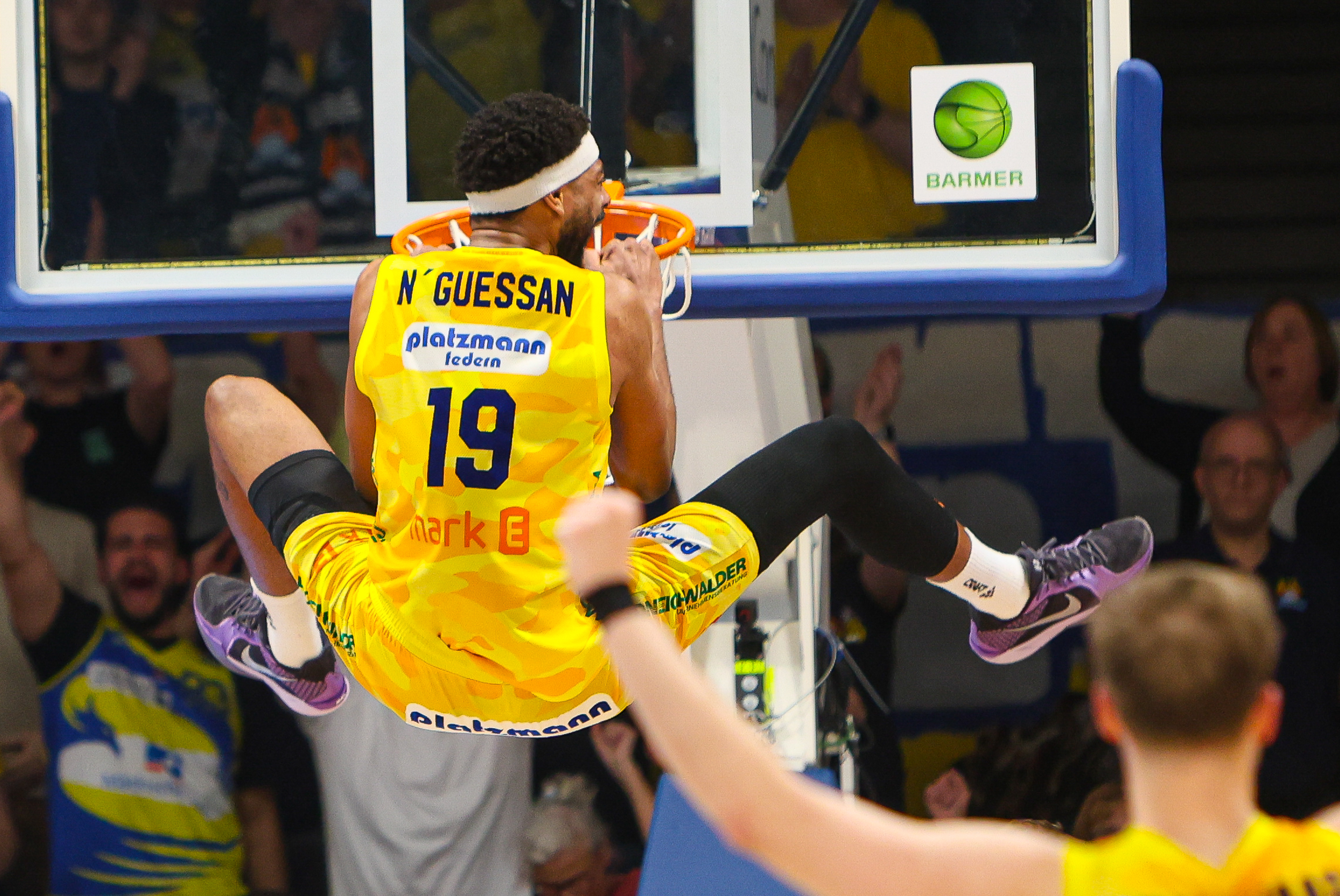 Ein Basketballspieler in gelbem Trikot hängt lachend am orangefarbenen Basketballkorb