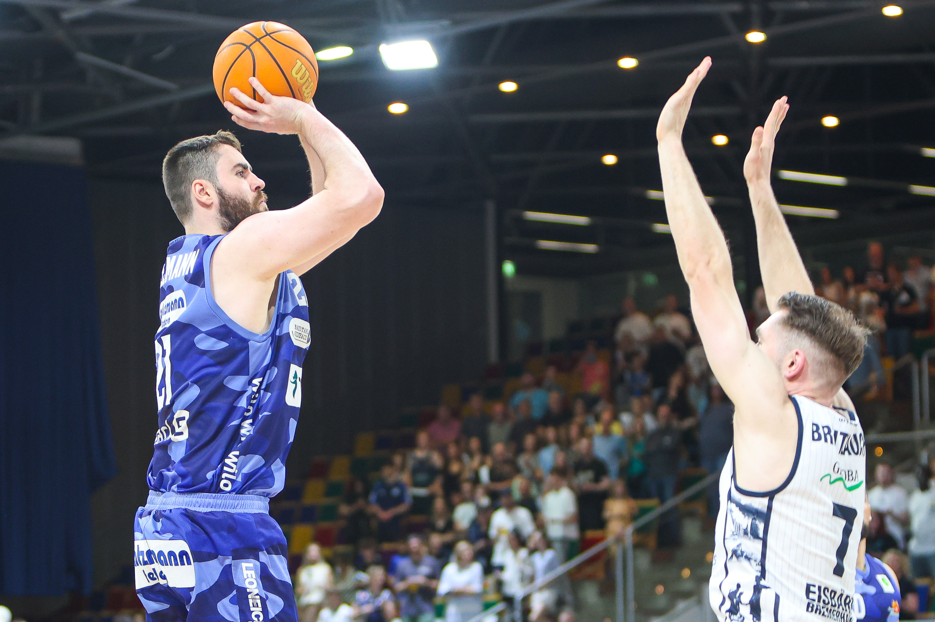 Ein Basketballspieler in blauem Trikot wirft konzentriert den Ball, Gegenspieler versucht zu blocken