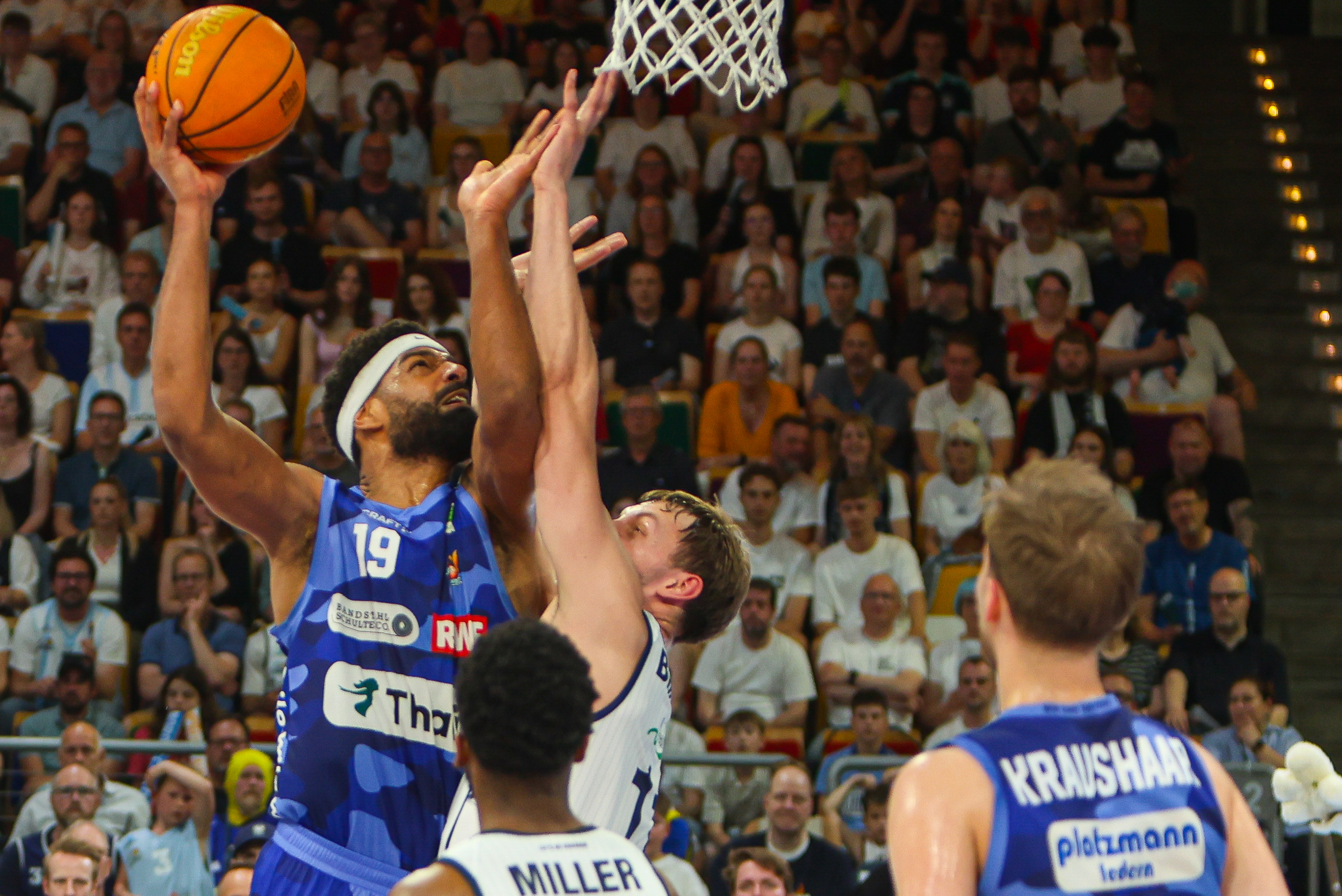 Ein Basketballspieler springt konzentriert zum Korb, blaue Trikots und volle Tribüne