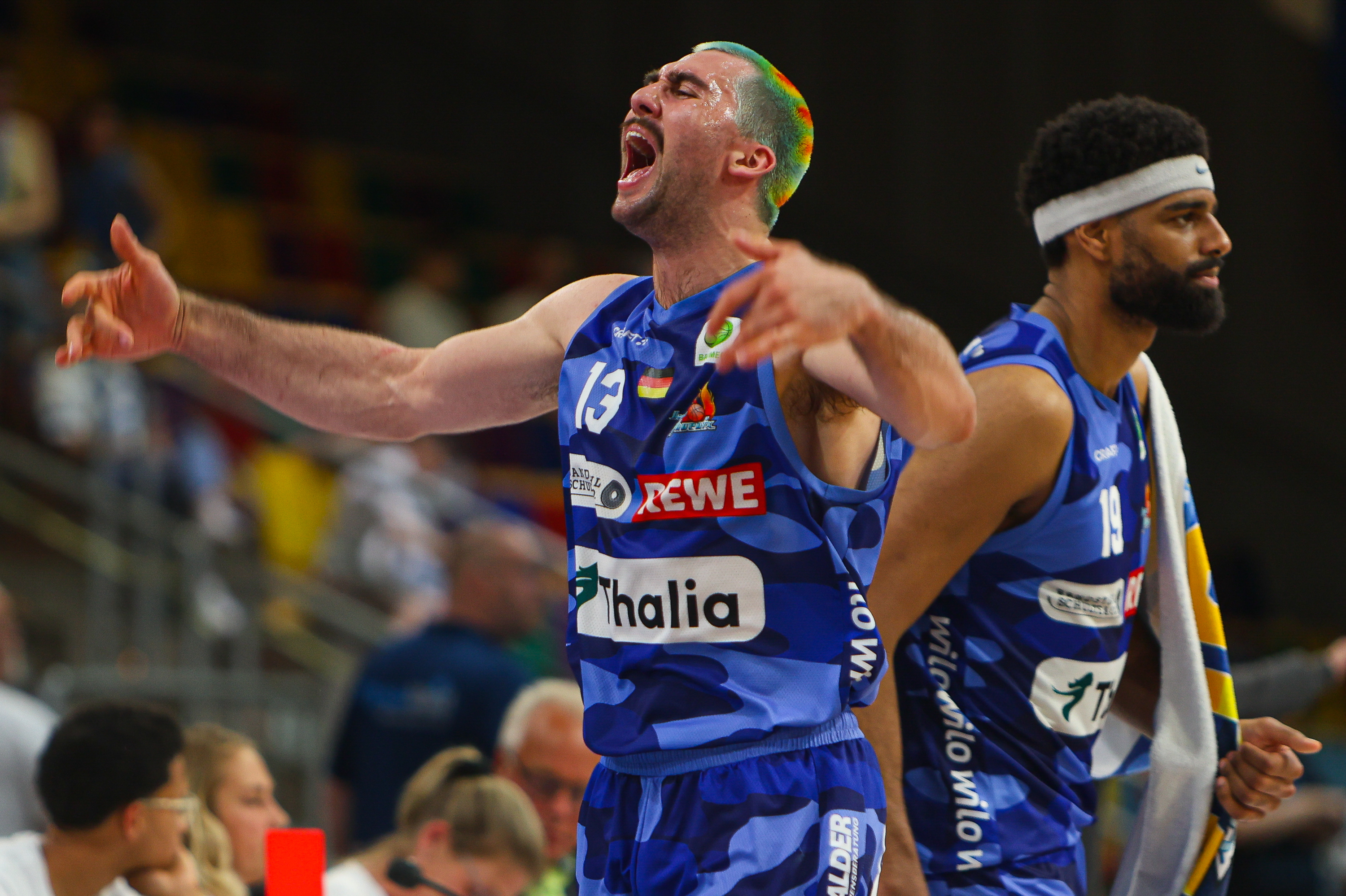 Ein Basketballspieler mit ernstem Blick trägt ein blaues Trikot und hebt die Arme