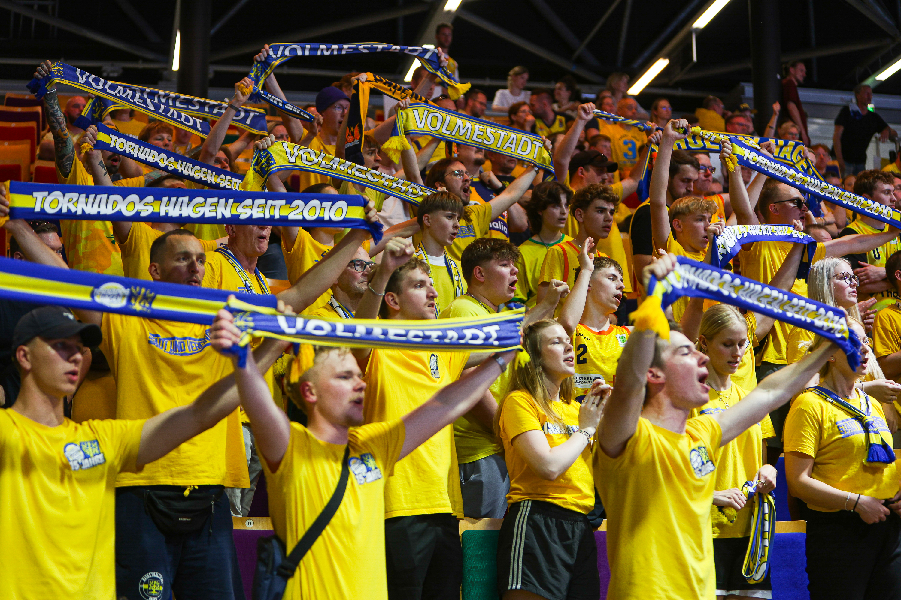 Eine Gruppe Fans mit gelben Shirts jubelt mit bunten Schals in einer Sportarena