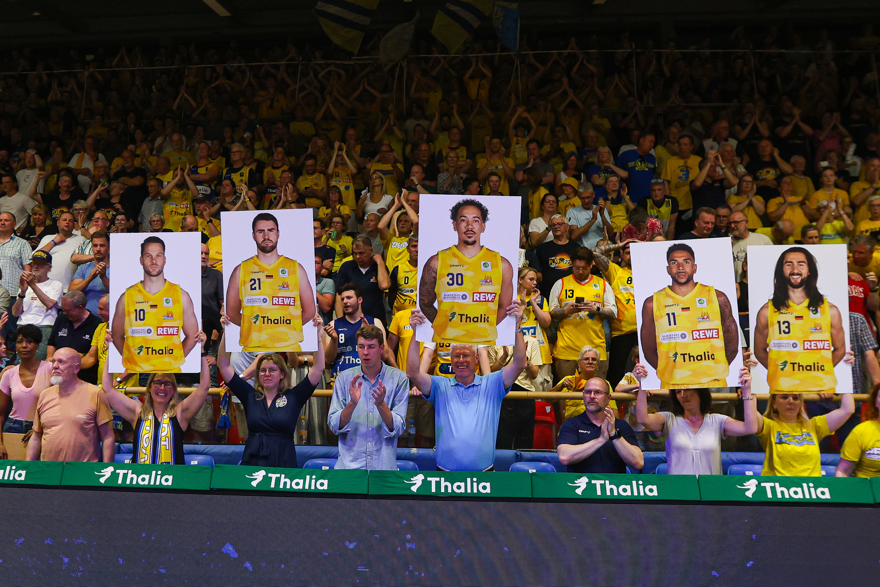 Jubelnde Fans in Gelb halten Spielerposter hoch, bunte Fahnen im Hintergrund