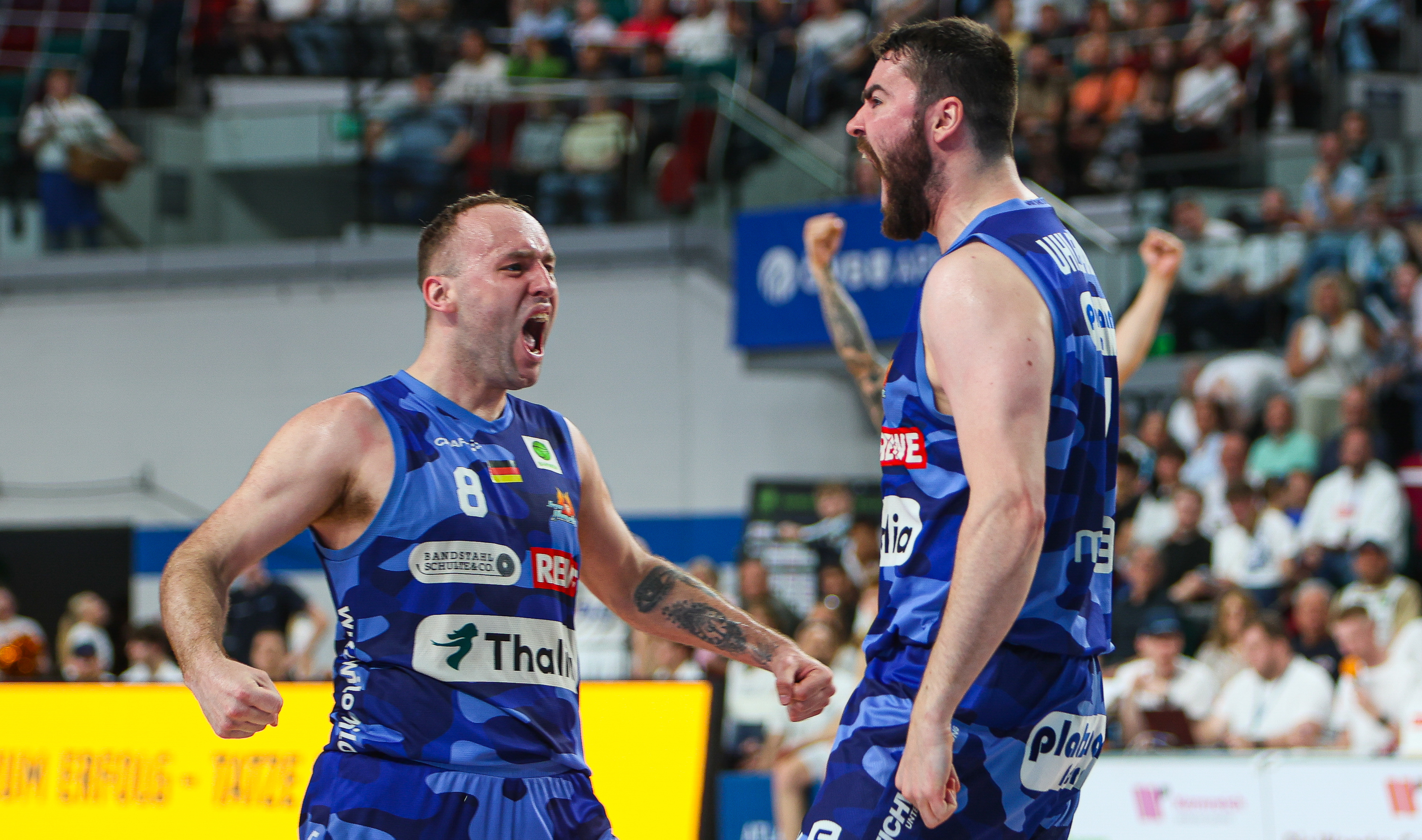 Zwei jubelnde Basketballspieler in blauen Trikots mit Tattoos im Stadion