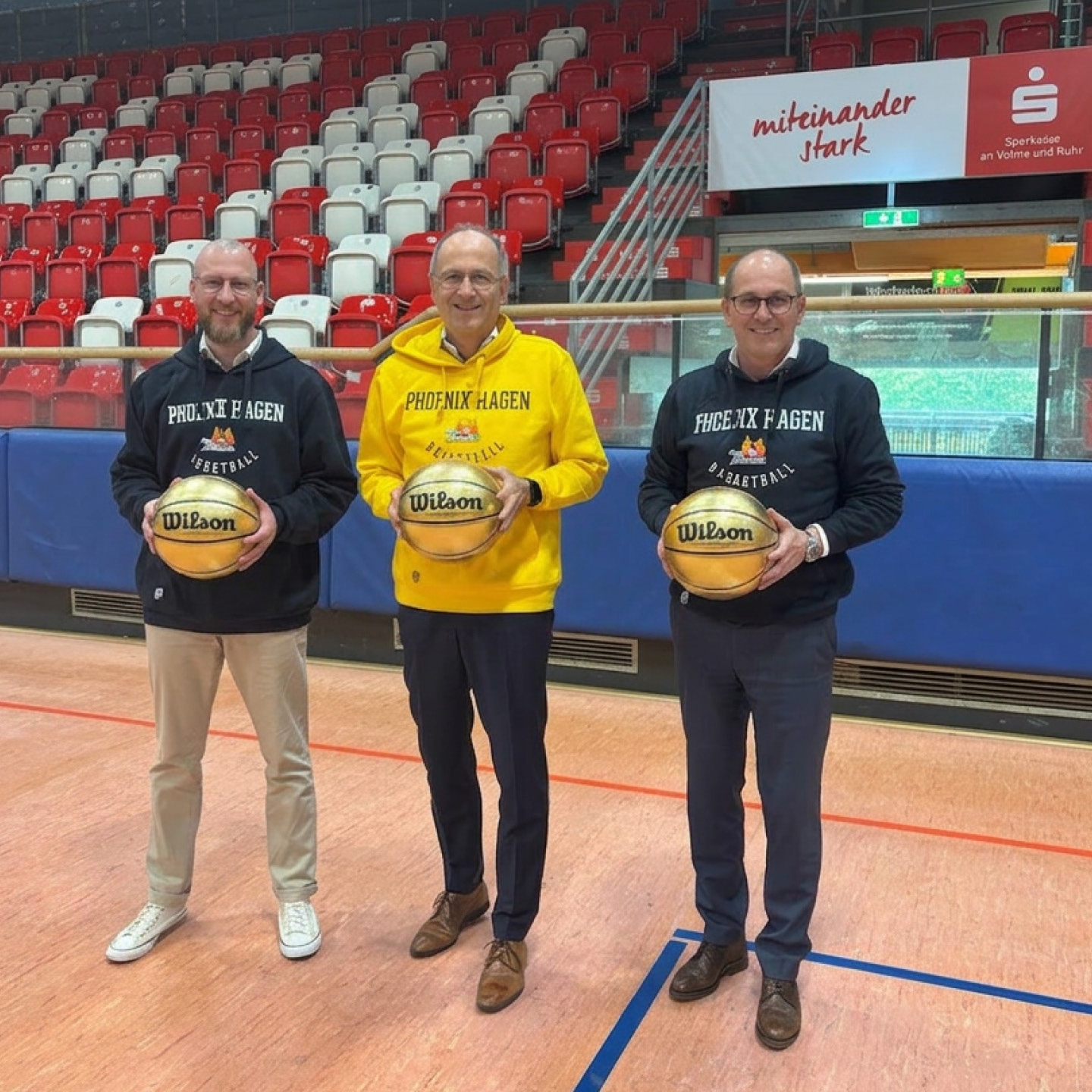 Drei ältere Männer lächeln in einer leeren Turnhalle und halten Phoenix Hagen Basketbälle
