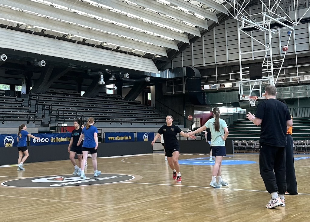 Mehrere junge Menschen spielen Basketball in einer großen Halle mit hellem Holzboden