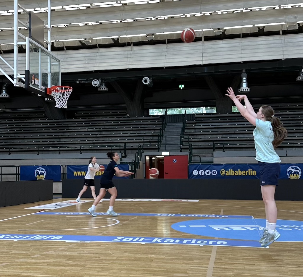 Eine junge Frau mit Zopf wirft einen Basketball, zwei andere laufen Richtung Korb
