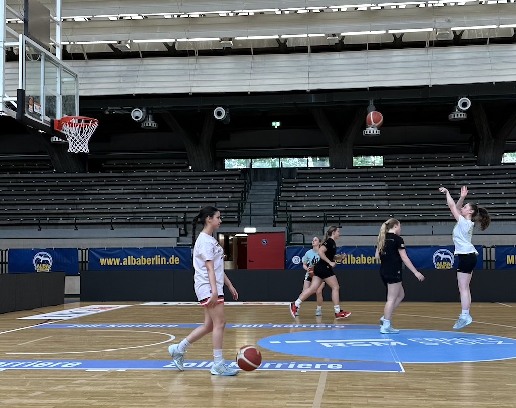 Fünf junge Frauen spielen Basketball in einer Halle, eine wirft den Ball zum Korb
