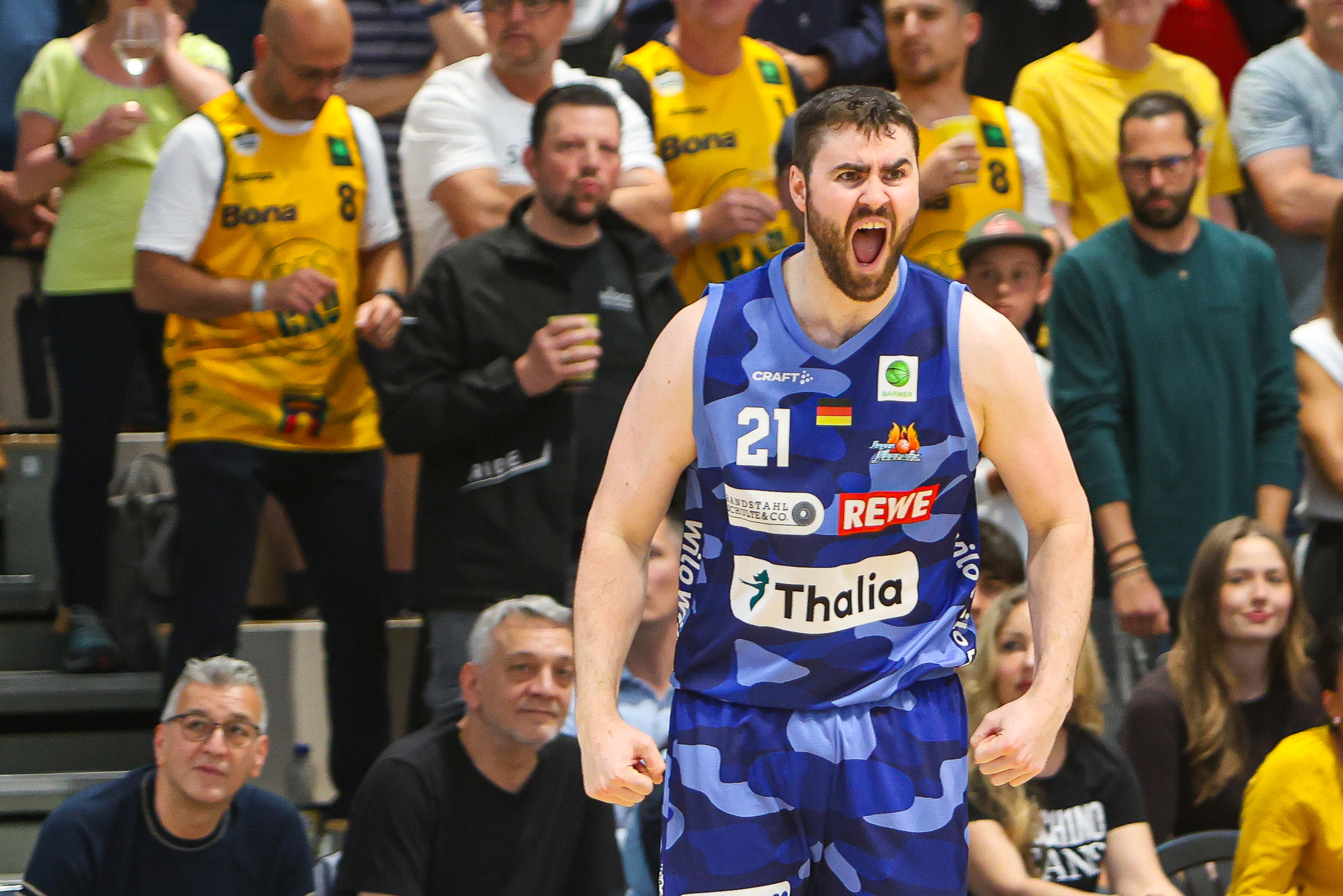 Entschlossener Basketballspieler im blauen Trikot 21 vor gelben Fans, Logos sichtbar