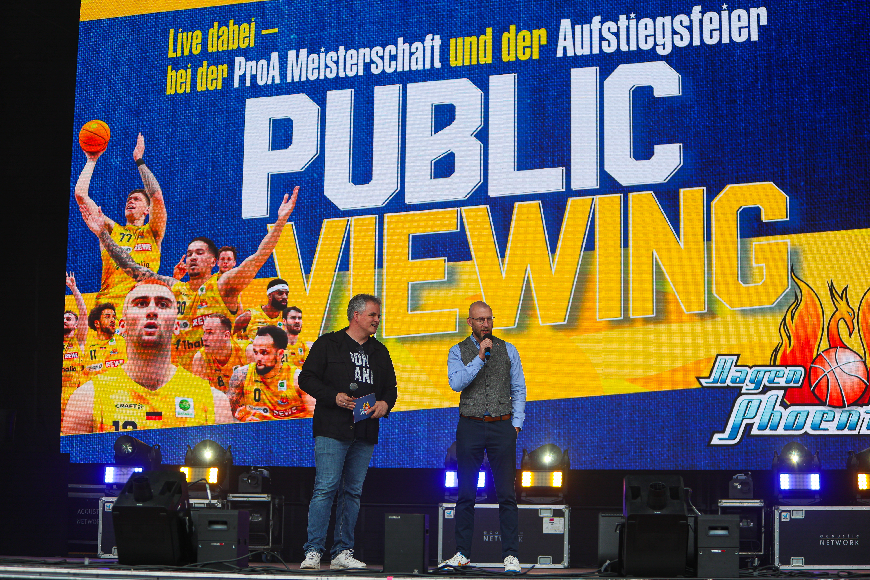 Zwei Maenner mit Mikrofon, freundlich, vor blau gelbem Public Viewing Banner