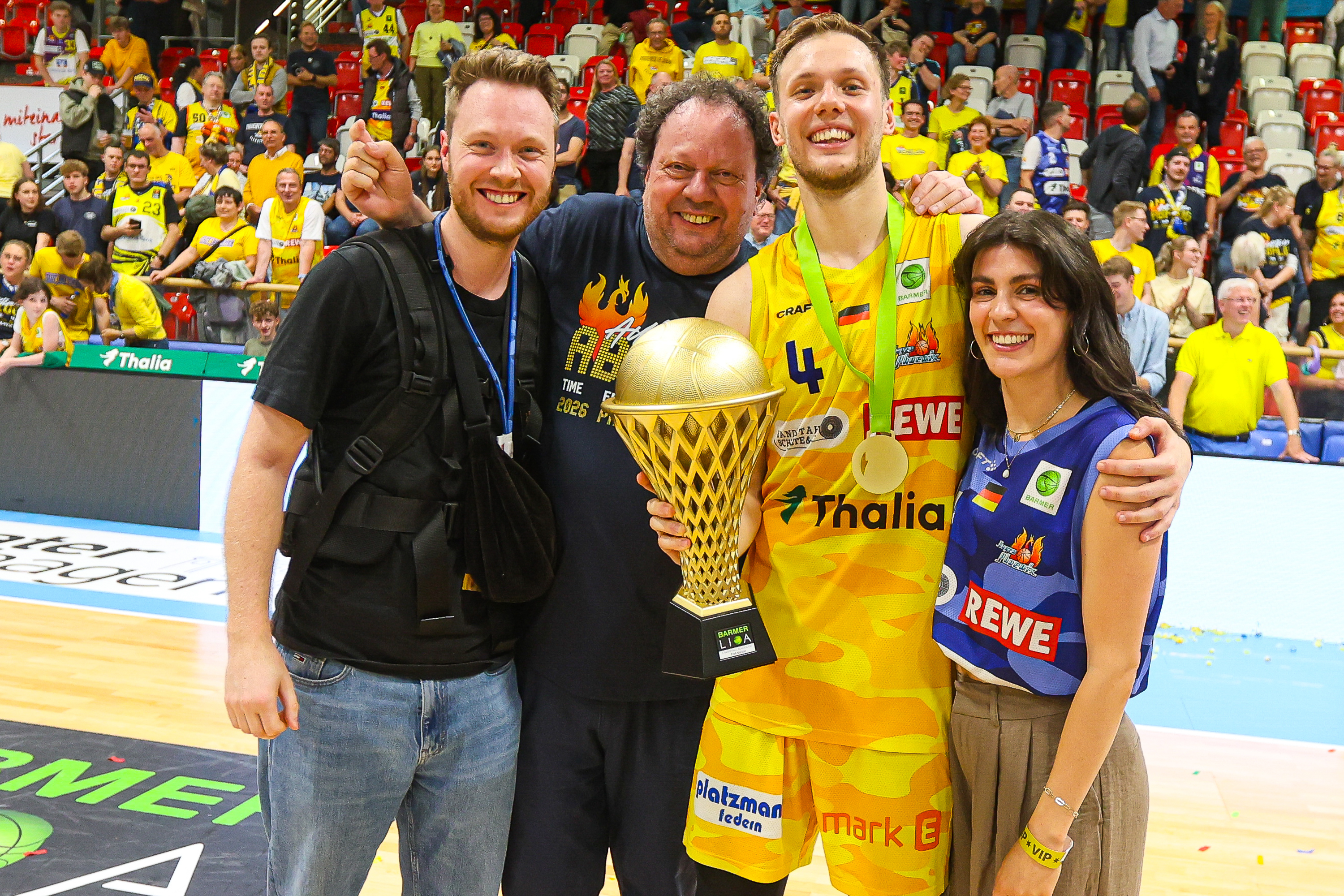 Vier Personen laecheln in Sporthalle mit goldenem Pokal und gelben Fans