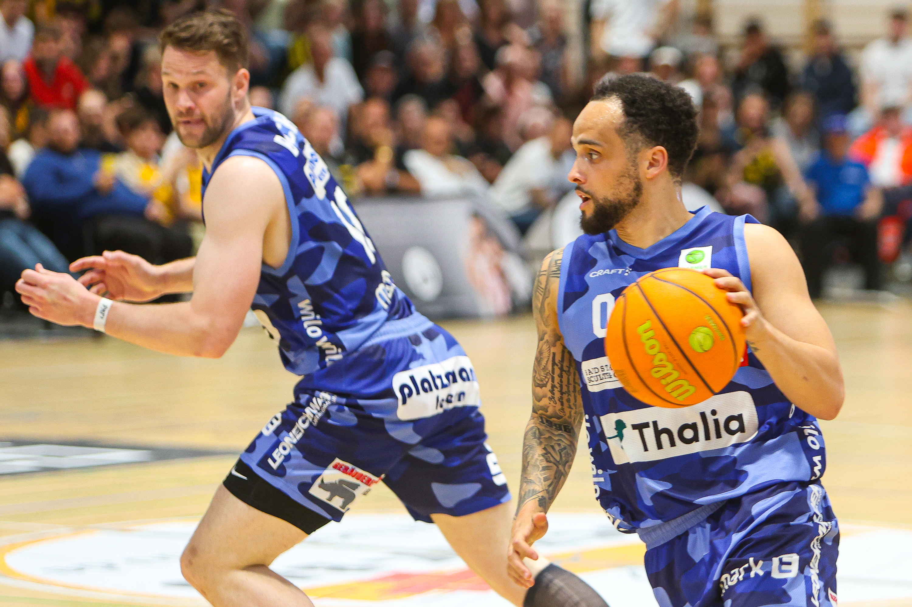 Zwei Basketballspieler in Blau, fokussiert, einer dribbelt orangen Ball, Tattoos
