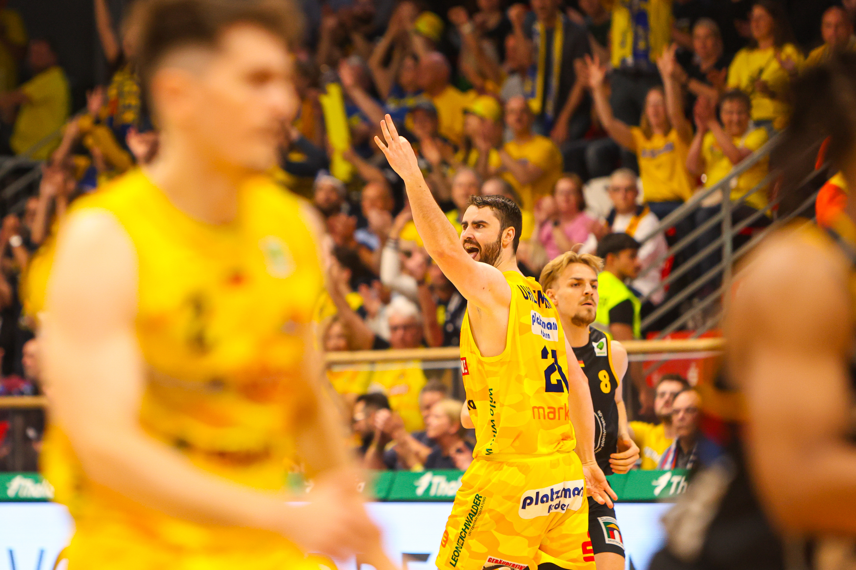 Jubelnder Basketballspieler im gelben Trikot, Arm erhoben, Halle gelb bunt