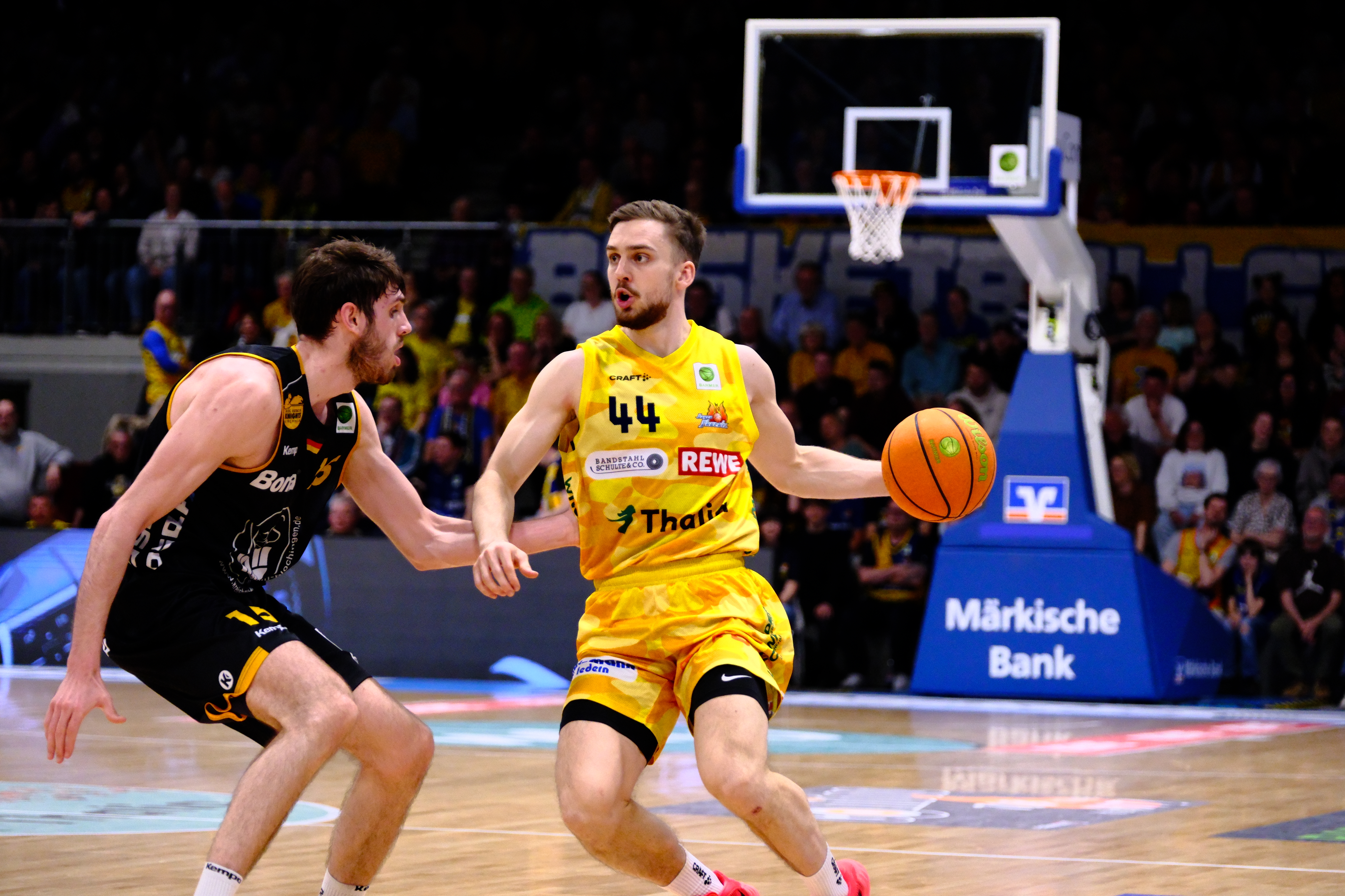 Fokussierter Basketballspieler in Gelb dribbelt, Gegner in Schwarz, Korb im Hintergrund