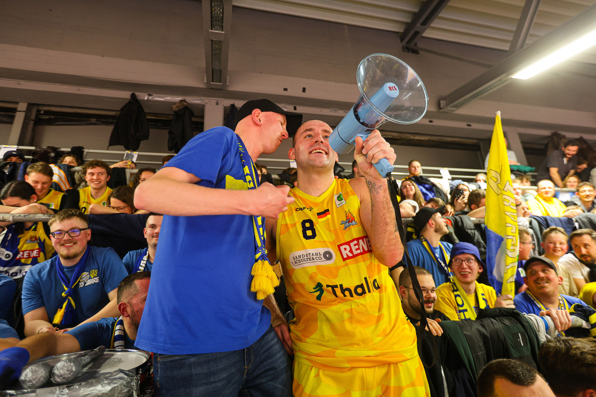 Zwei Fans laecheln, blau und gelb, einer mit Megafon, Fahnen im Hintergrund