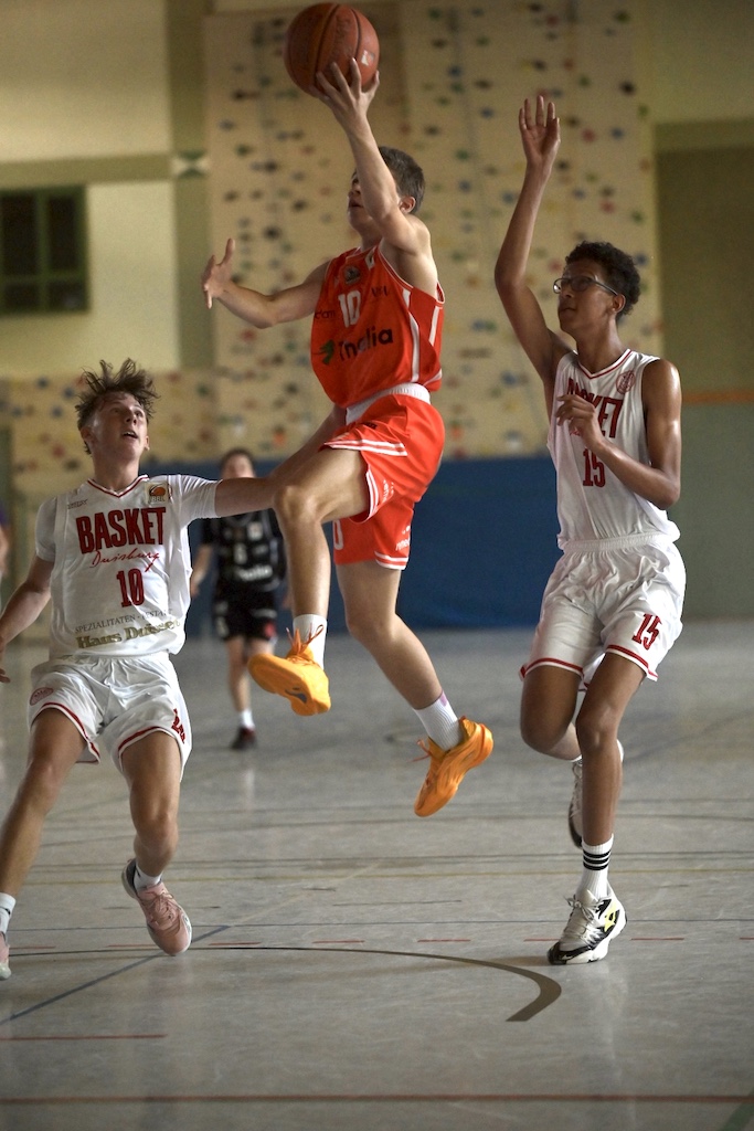Springender Basketballspieler in Rot, fokussiert, Ball hoch, Gegner in Weiss