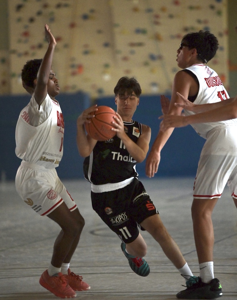 Fokussierter Basketballspieler in Schwarz dribbelt zwischen zwei Verteidigern in Weiss