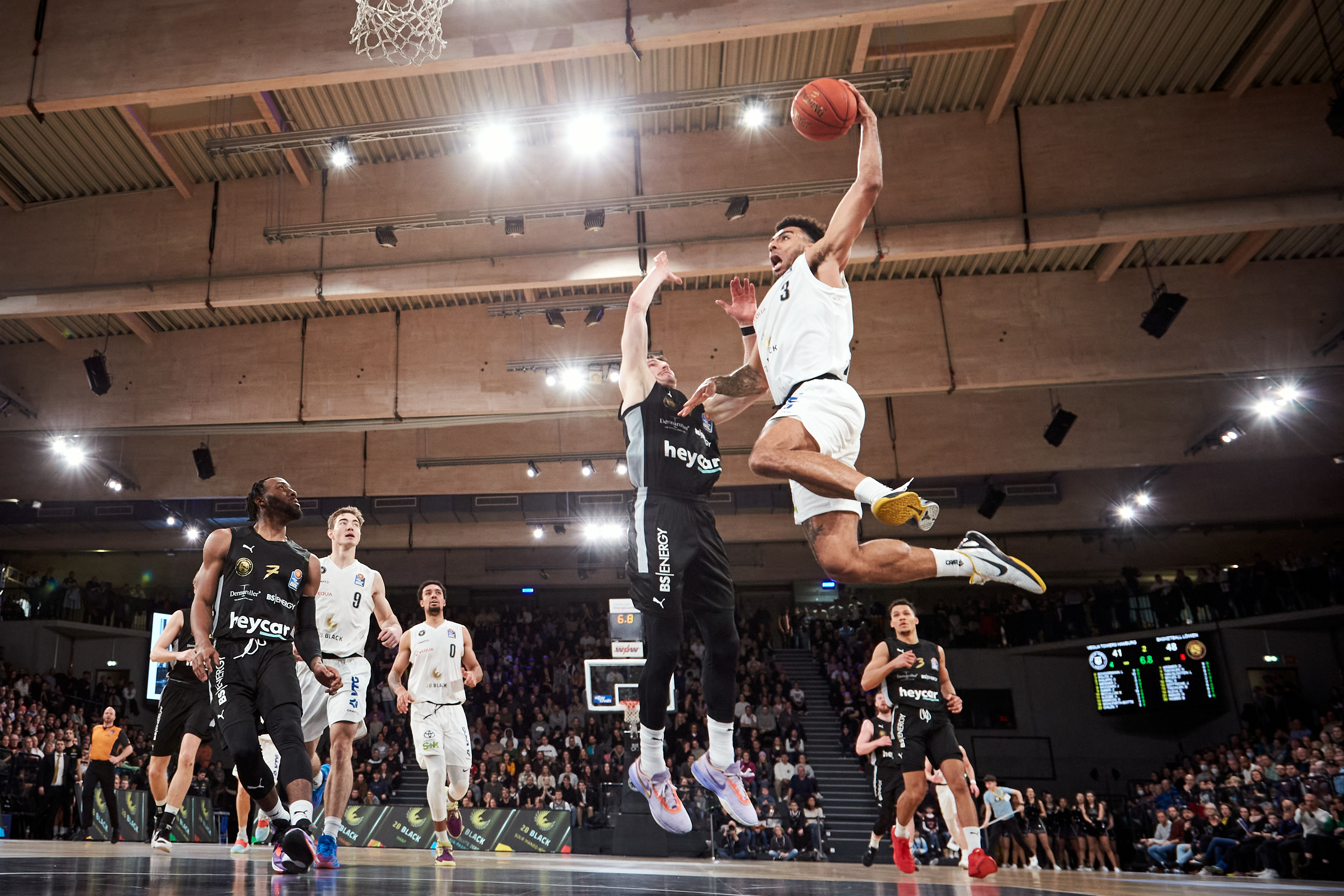 Springender Basketballspieler in Weiss laechelt beim Dunk ueber Gegner in Schwarz