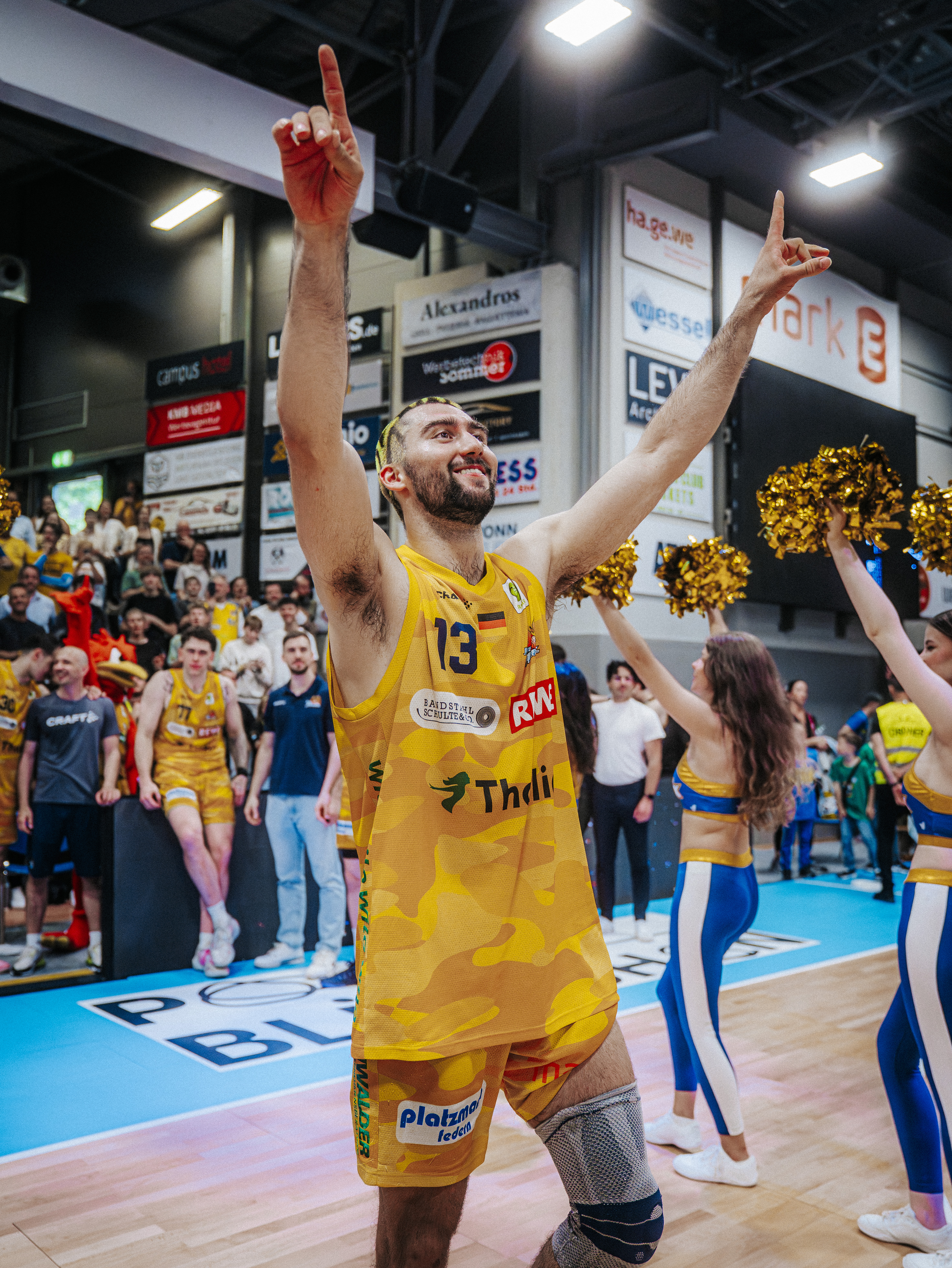 Jubelnder Basketballspieler im gelben Trikot hebt Arme, Fans dahinter