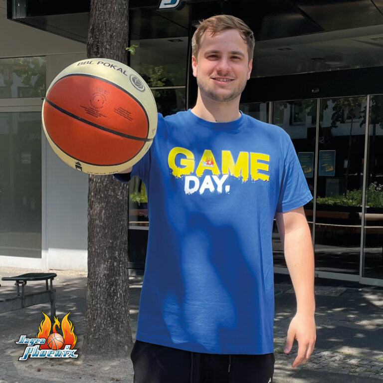 Laechelnder Mann in blauem GAME DAY Shirt haelt orange Basketball hoch