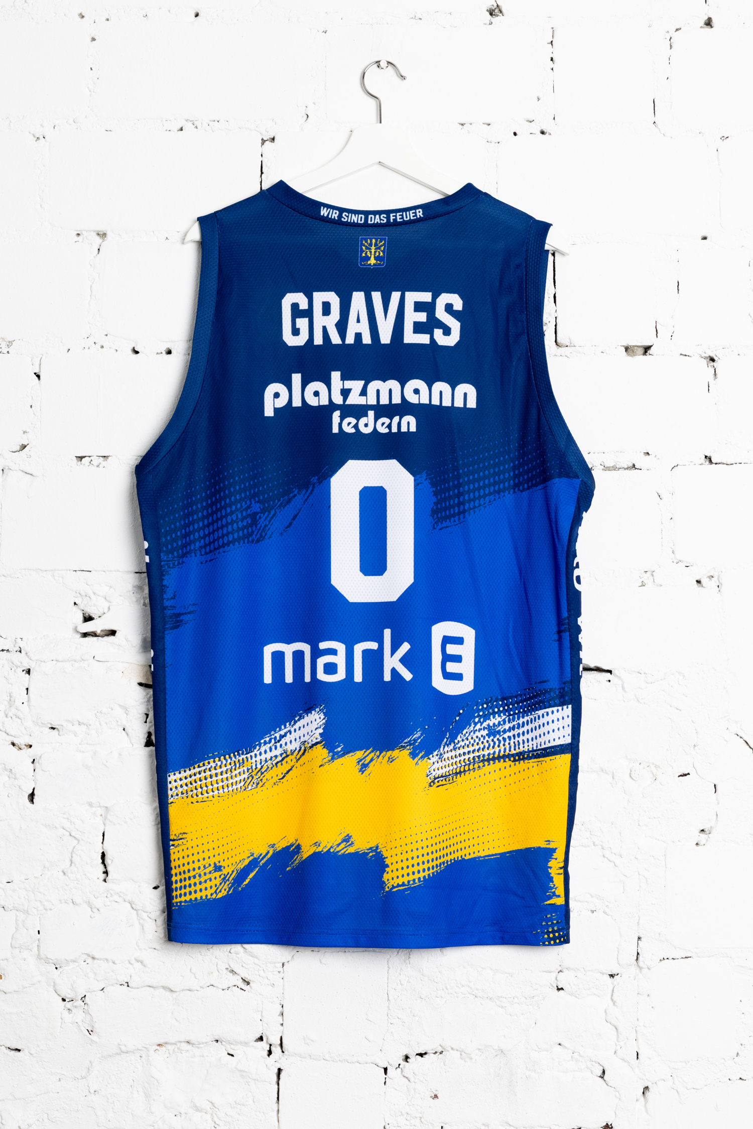 Blaugelbes Trikot mit Aufdruck GRAVES 0 und Mark E an weißer Wand hängend