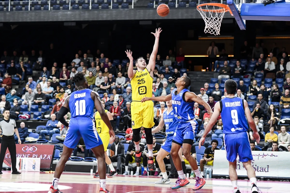 Konzentrierter Basketballspieler in Gelb beim Sprungwurf, Gegner in Blau, orangefarbener Ball am Korb