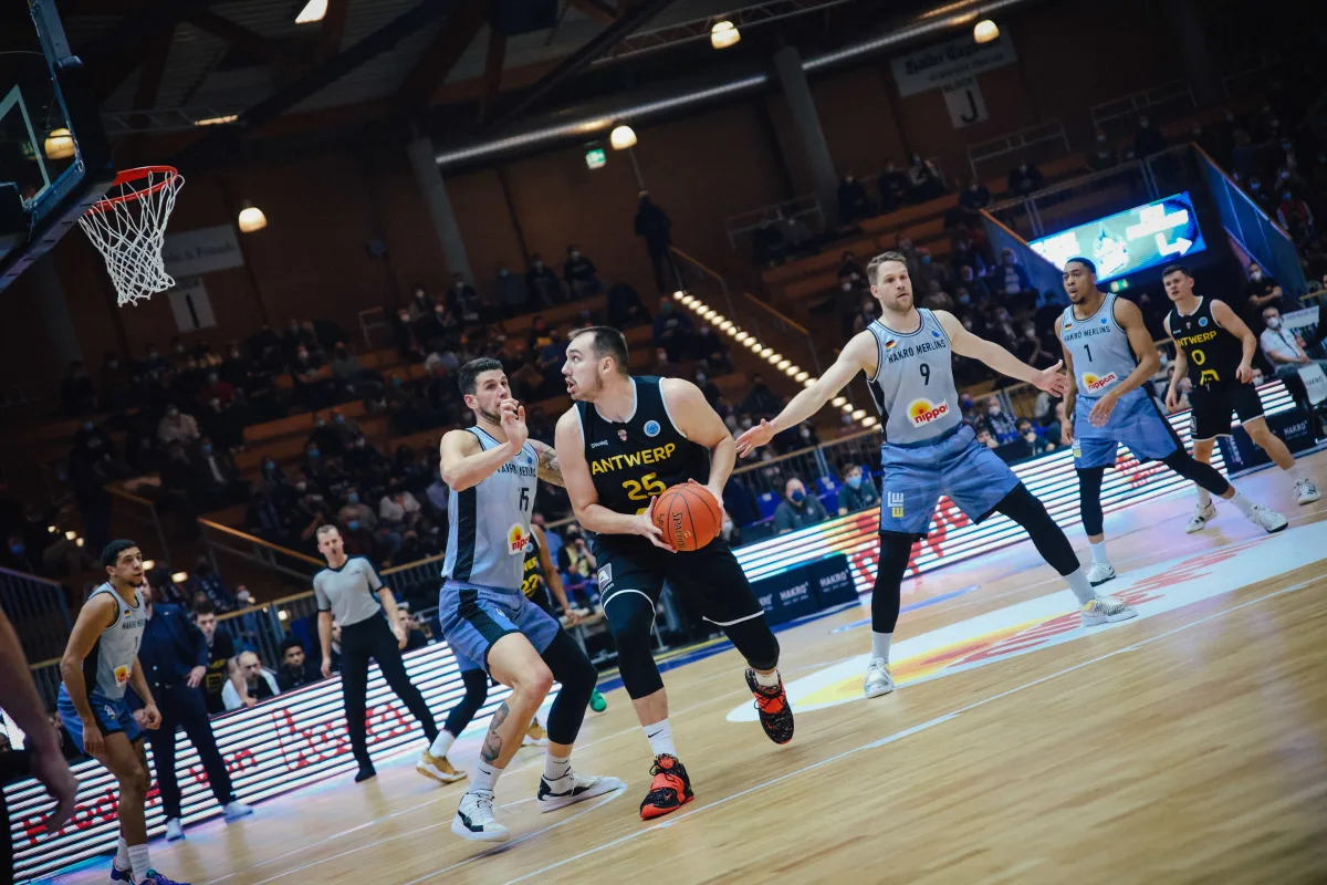 Konzentrierter Basketballspieler in schwarzem Trikot mit orangefarbenem Ball, umringt von Verteidigern in hellblauen Trikots, in heller Halle mit Zuschauern und Korb links oben