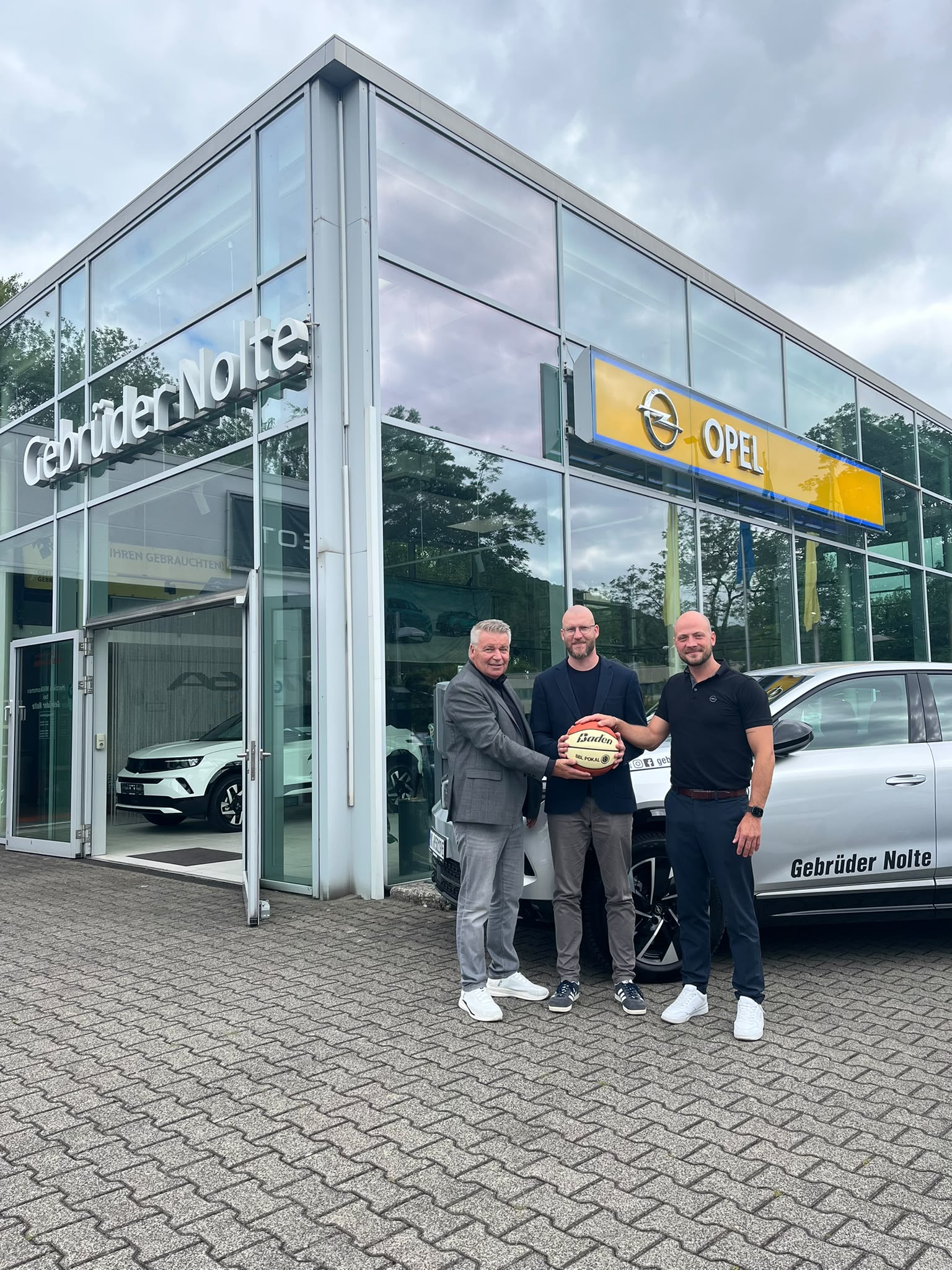Drei Maenner laecheln vor Glas Autohaus, Grau Blau, mit Ball und Opel Schild