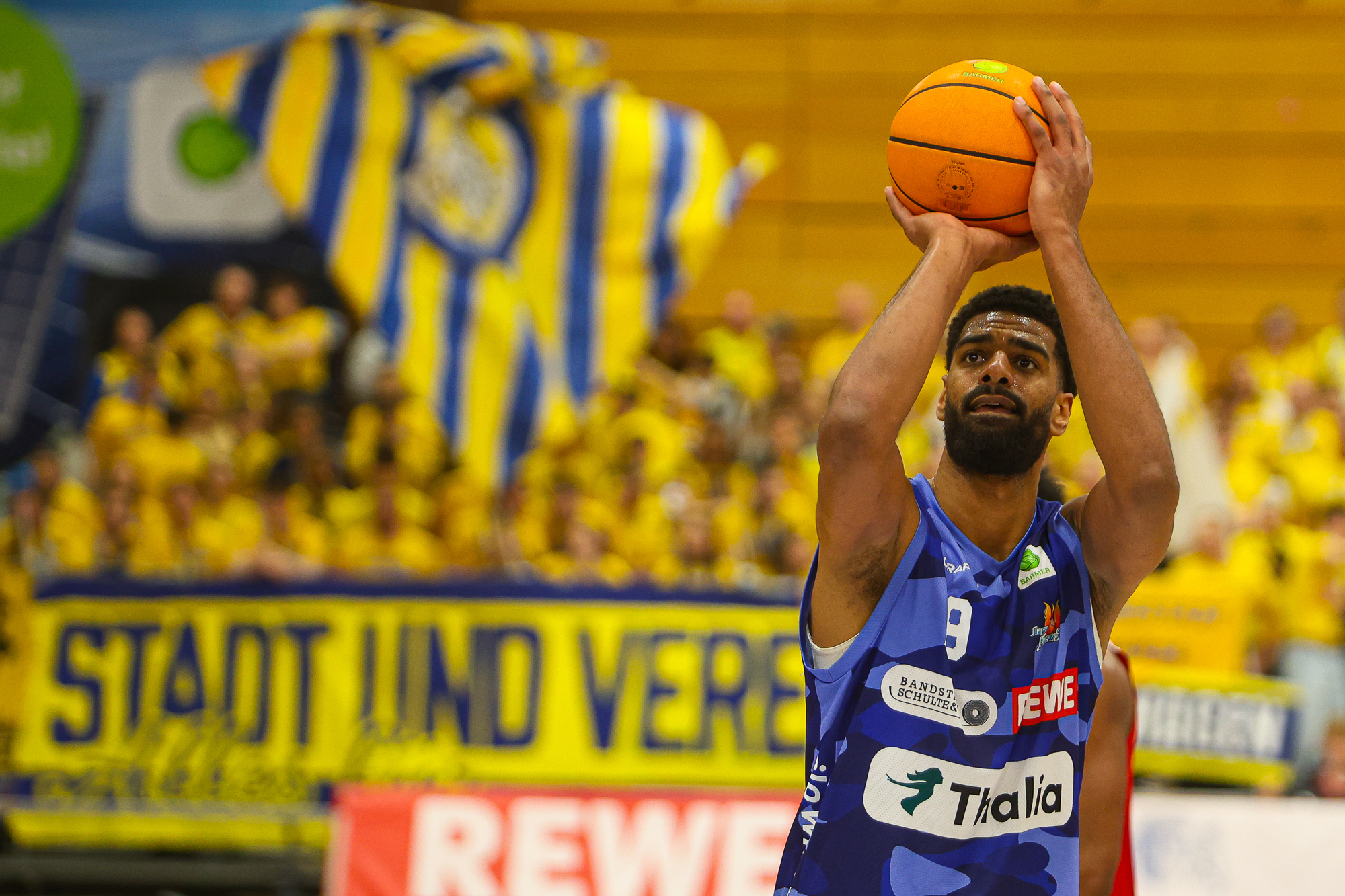 Konzentrierter Mann im blauen Trikot wirft orangen Basketball, gelbe Fans im Hintergrund