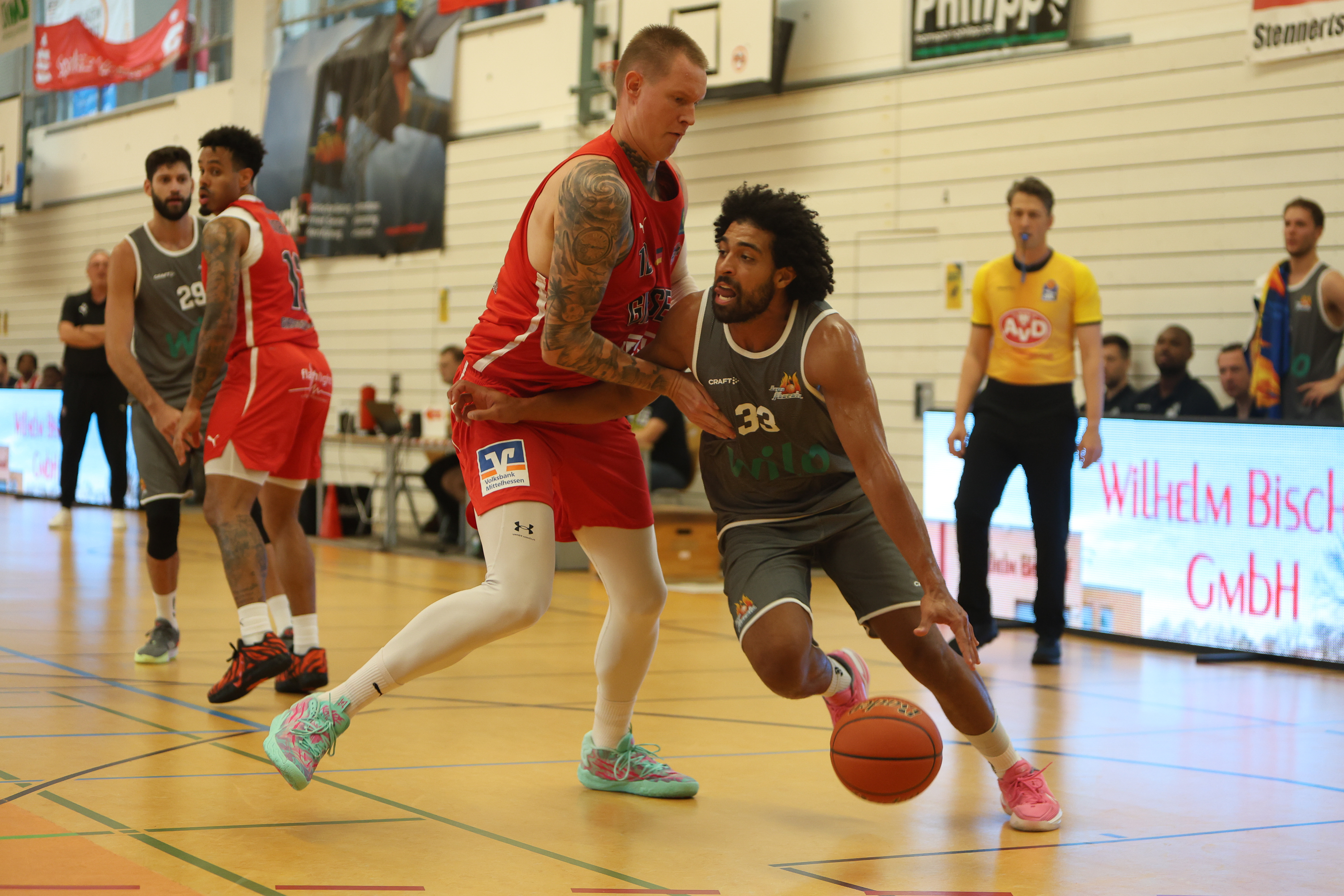 Basketballspieler in grauem Trikot dribbelt konzentriert am Gegner in Rot vorbei