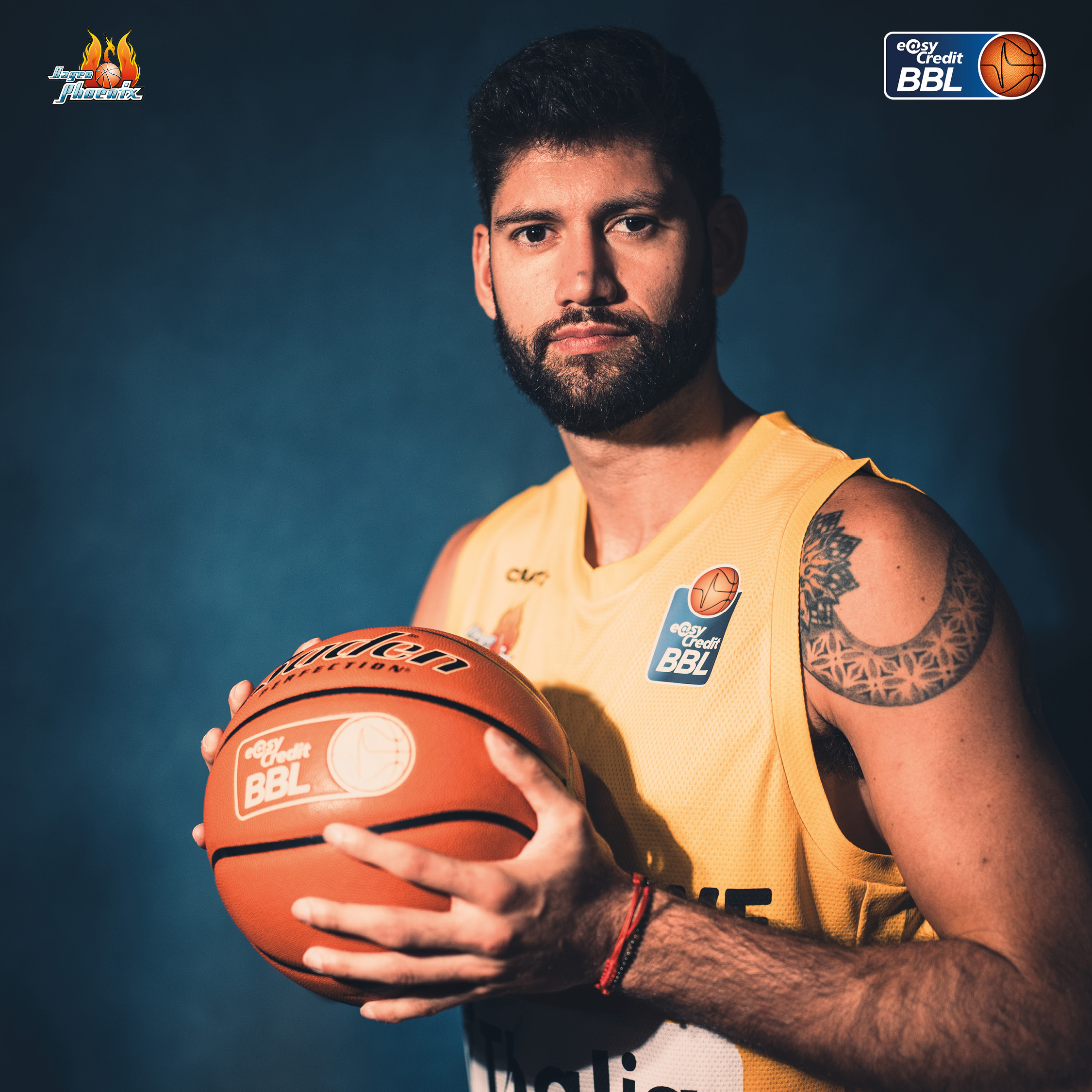 Ernster Basketballspieler mit Bart und Tattoo im gelben Trikot haelt einen Ball