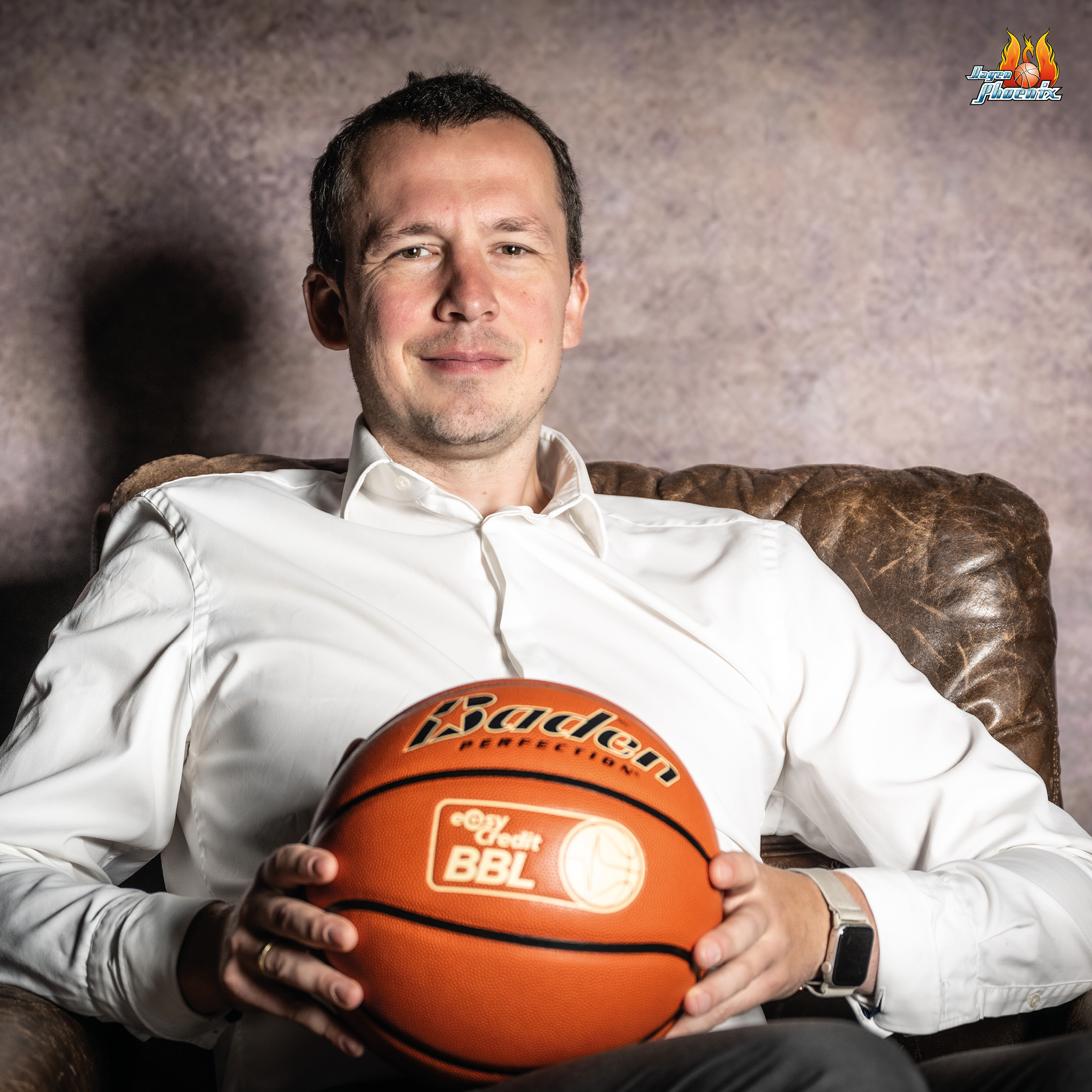 Ein laechelnder Mann im weissen Hemd haelt einen orangefarbenen Basketball