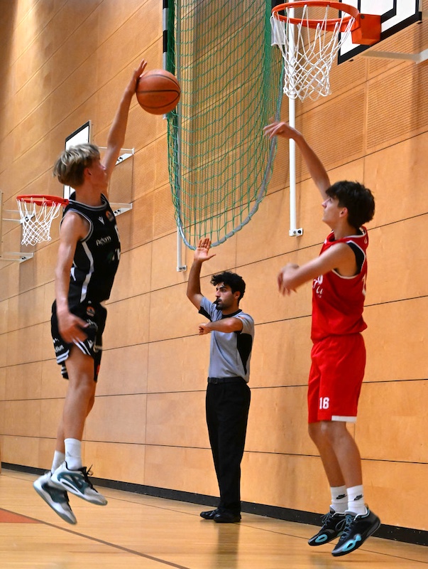 Zwei Basketballspieler springen konzentriert waehrend ein Schiedsrichter zusieht