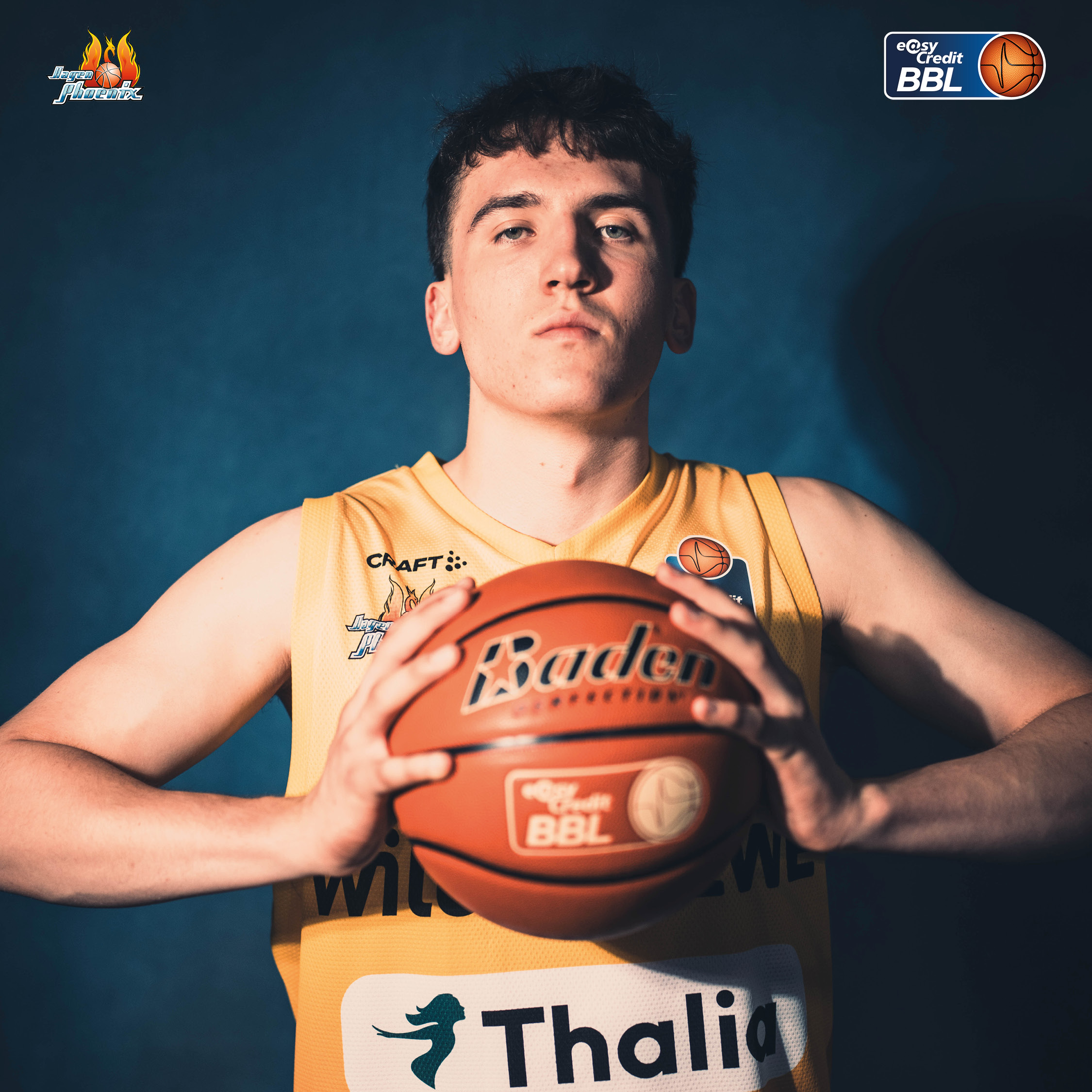 Junger Basketballspieler im gelben Trikot haelt ernst einen Ball vor blauem Hintergrund