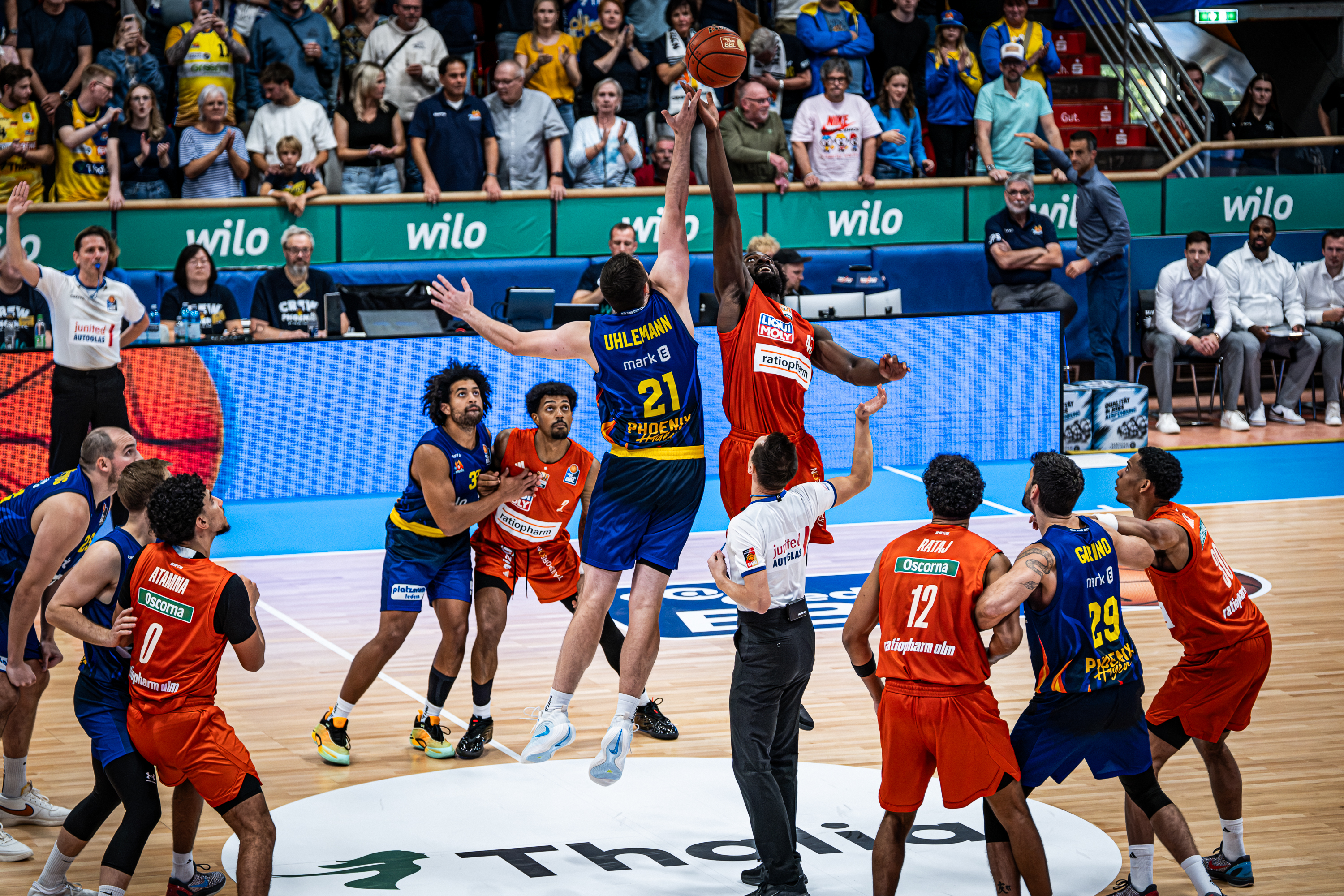 Zwei Basketballspieler in Blau und Orange springen konzentriert zum Sprungball