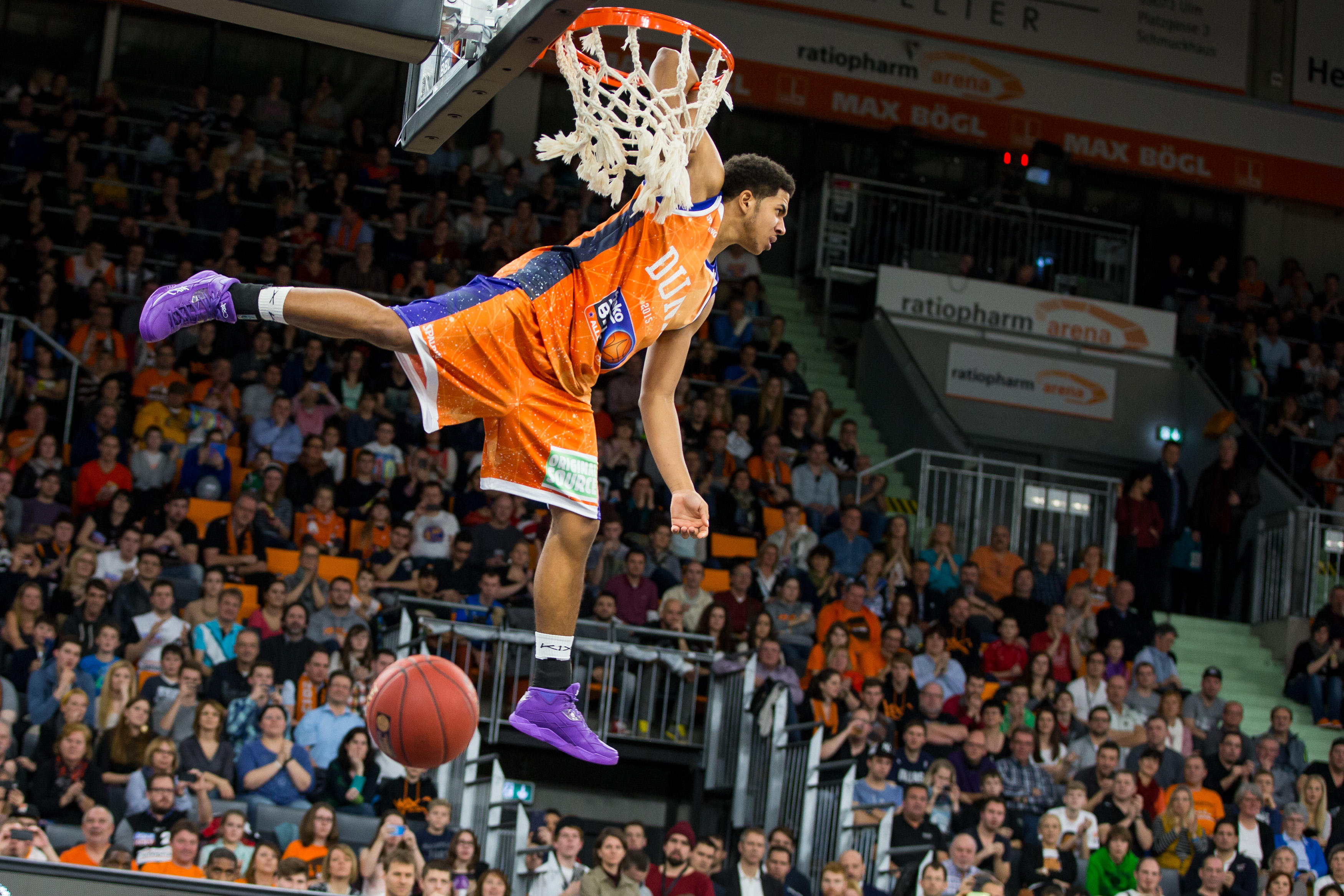 Basketballspieler in orangem Trikot haengt konzentriert am Korb ueber Publikum