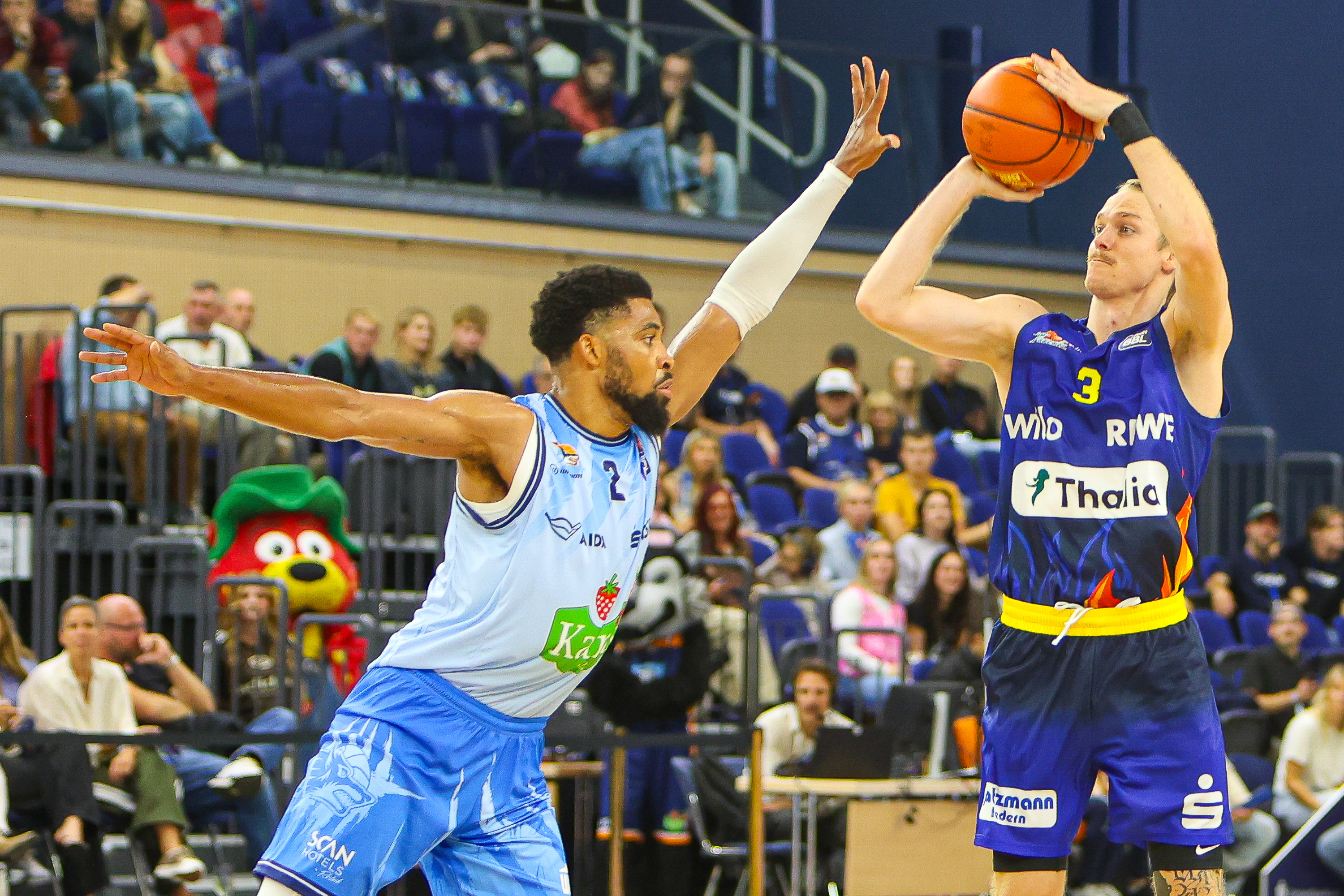 Ein Basketballspieler in Blau wirft den Ball waehrend ein Gegner ihn blockt