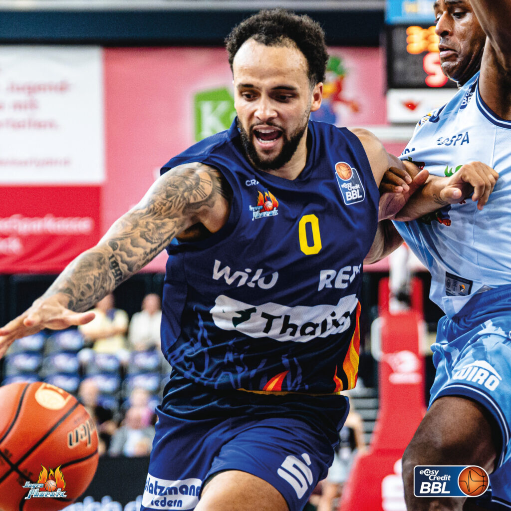 Basketballspieler mit blauem Trikot und Tattoos dribbelt konzentriert am Gegner vorbei