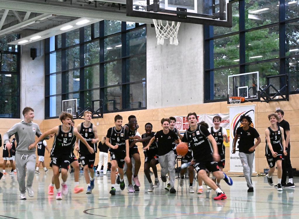 Jubelnde Basketballmannschaft in schwarzen Trikots rennt freudig ueber das Spielfeld