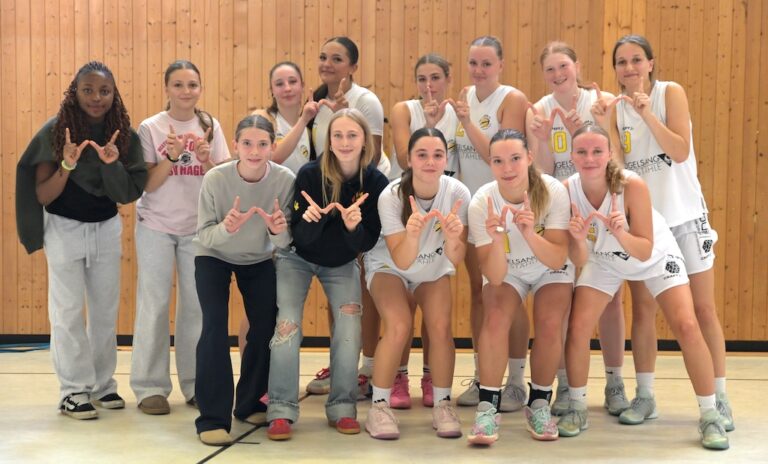 Laechelnde Basketballmannschaft in weissen Trikots formt Buchstaben mit Haenden