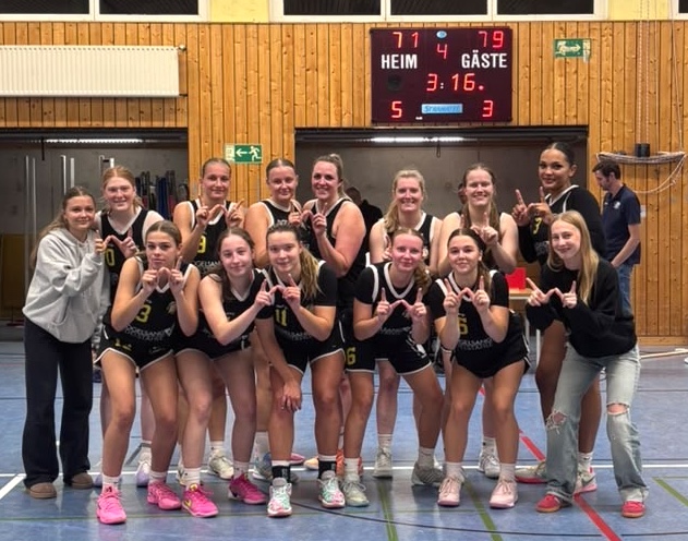 Ein laechelndes Basketballteam in schwarzen Trikots posiert vor der Anzeigetafel