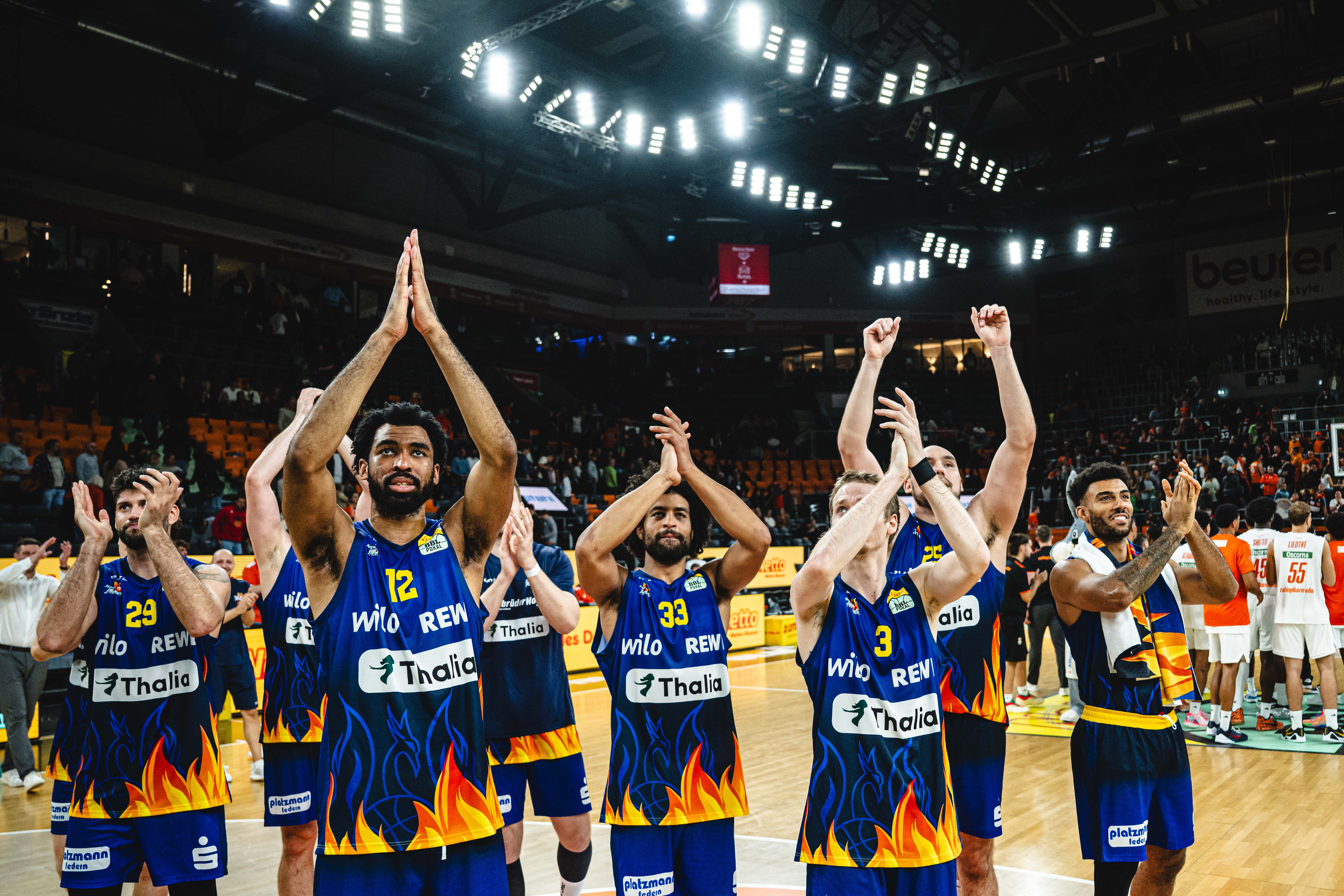 Basketballspieler in blauen Trikots mit Flammenmuster applaudieren jubelnd den Fans