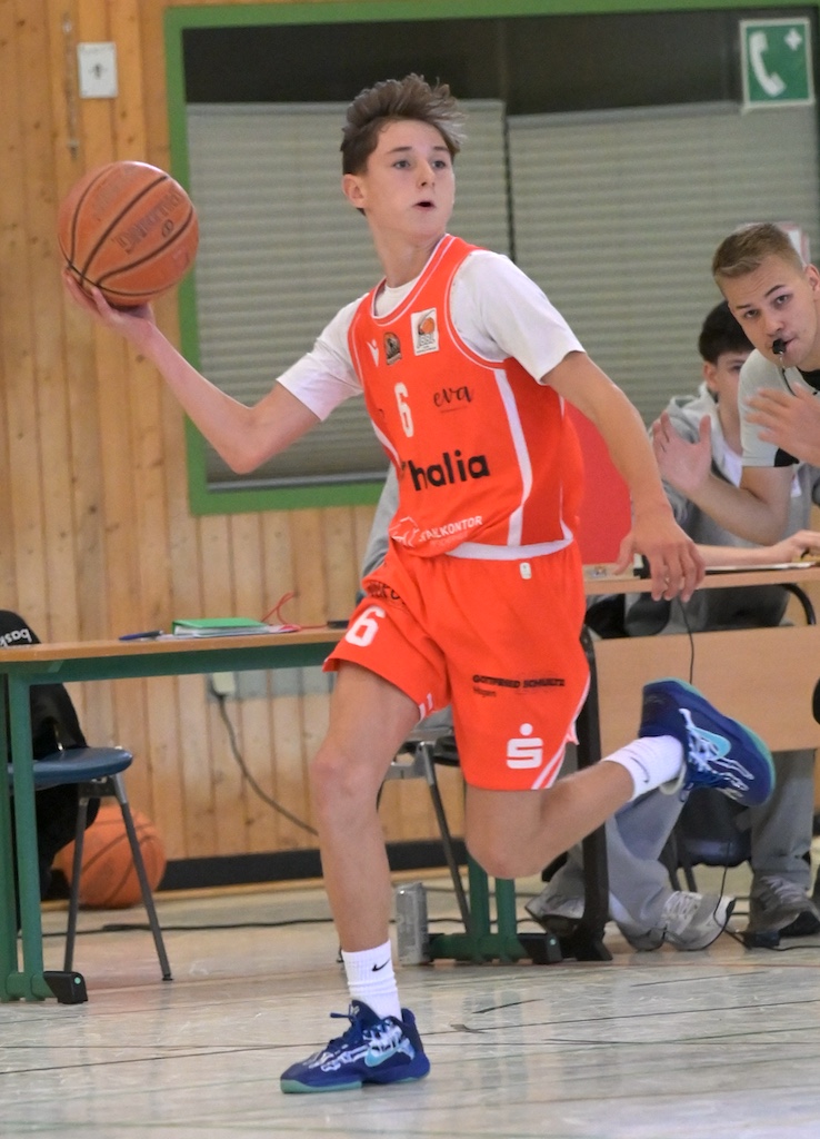 Ein junger Basketballspieler im orangefarbenen Trikot haelt konzentriert den Ball