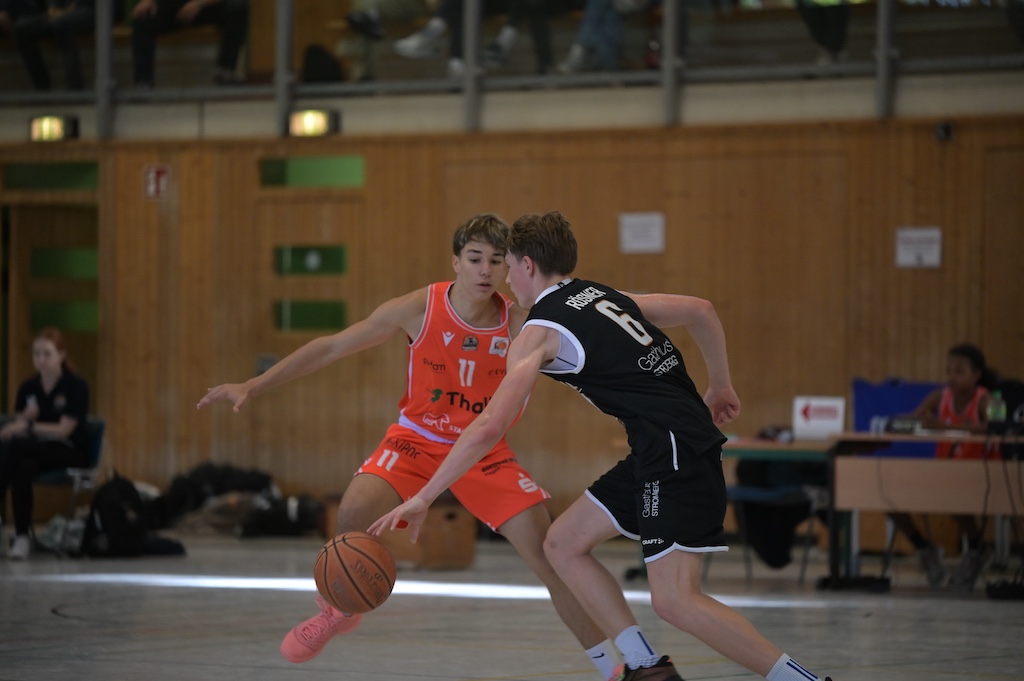 Zwei junge Basketballspieler in Orange und Schwarz kaempfen konzentriert um den Ball