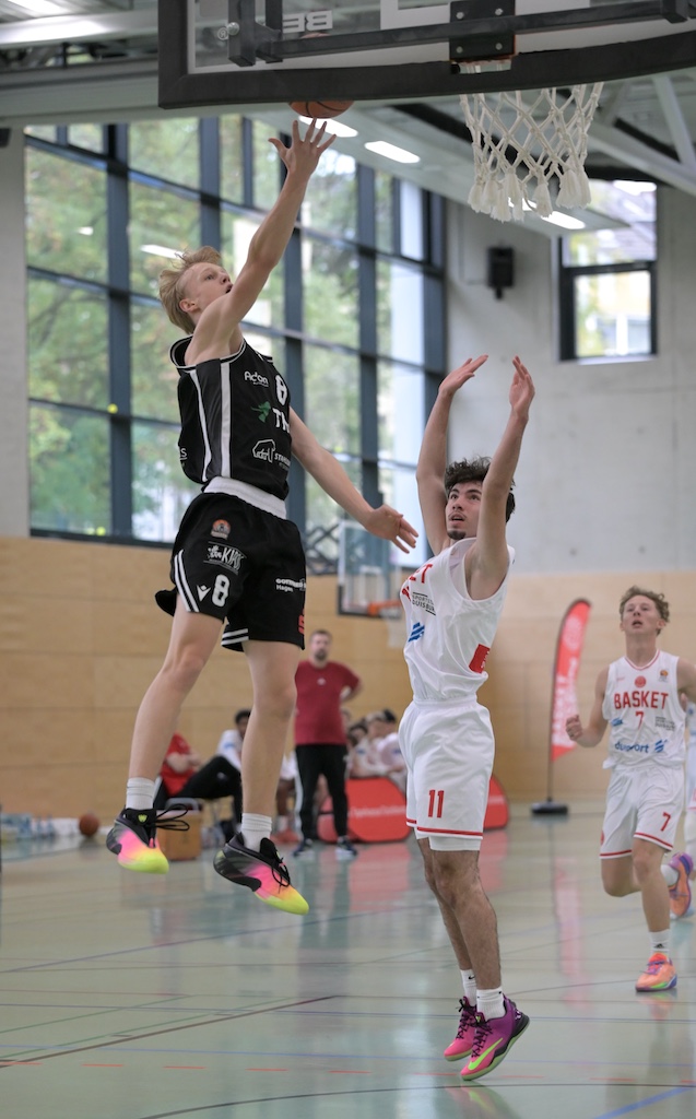 Ein Basketballspieler in schwarzem Trikot springt zum Korbwurf vor dem Gegner