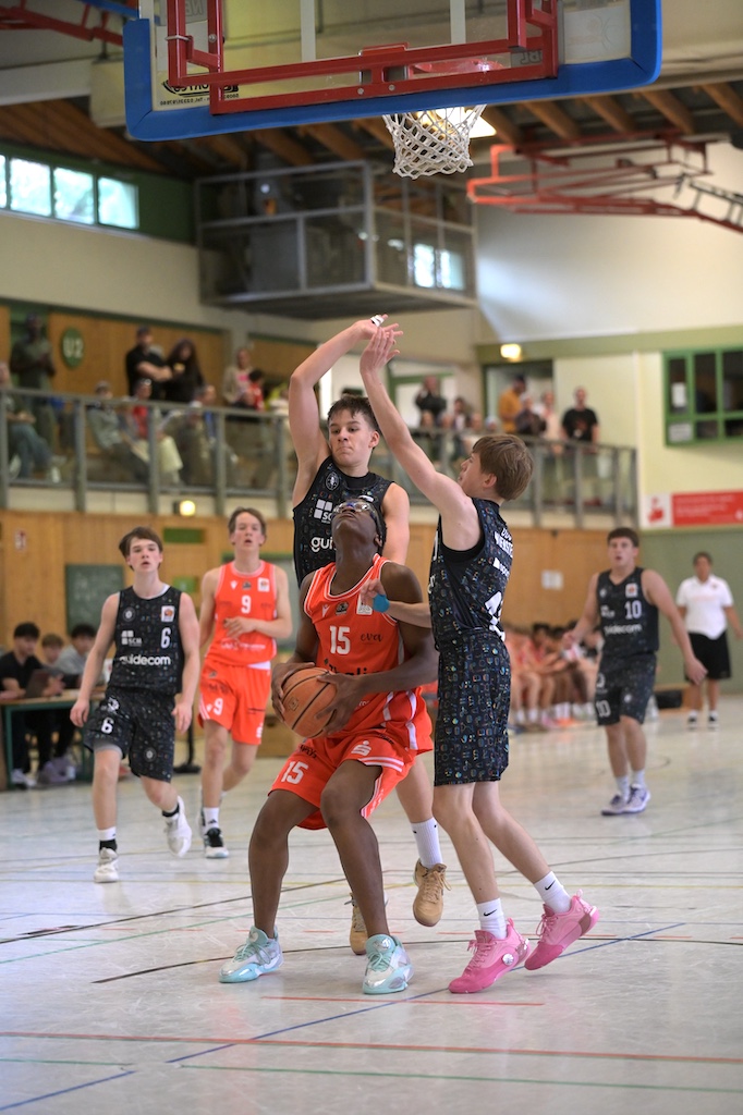Basketballspieler in orangem Trikot mit Nummer 15 wird von zwei Gegnern verteidigt