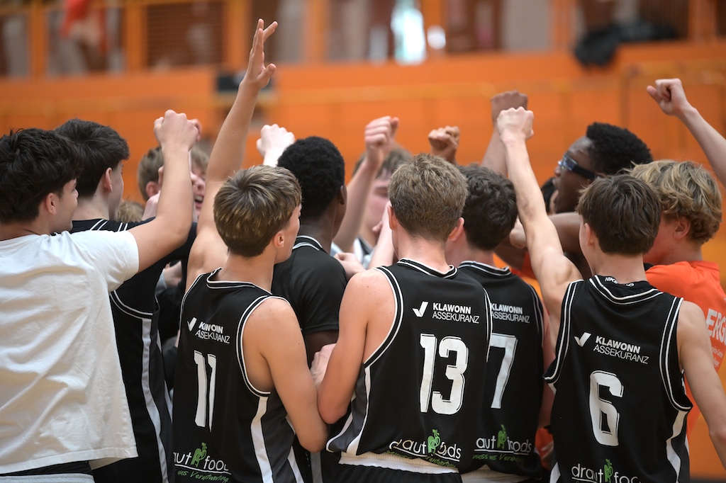 Ein Basketballteam in schwarzen Trikots feiert mit erhobenen Armen zusammen