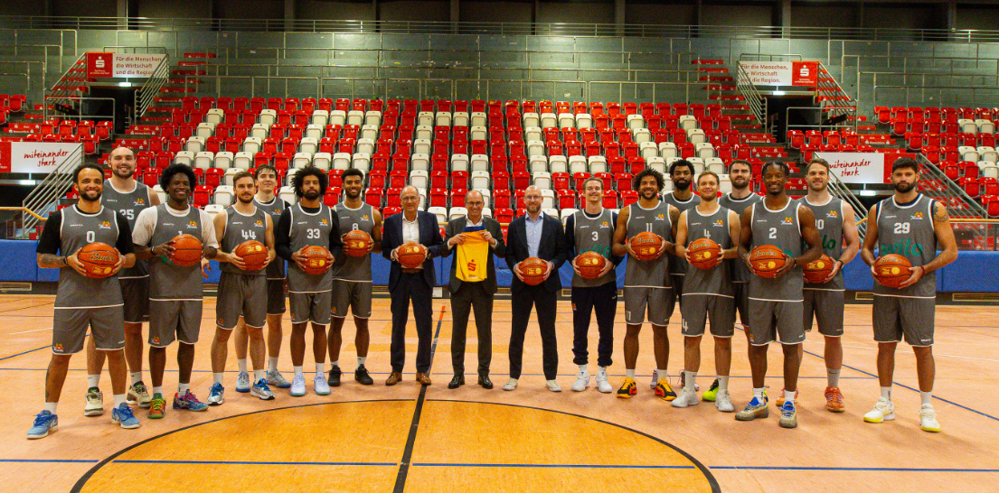 Basketballmannschaft in grauen Trikots posiert mit Baellen in einer Halle