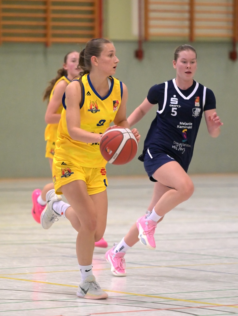 Basketballspielerin in gelbem Trikot fuehrt konzentriert den Ball gegen Gegnerin